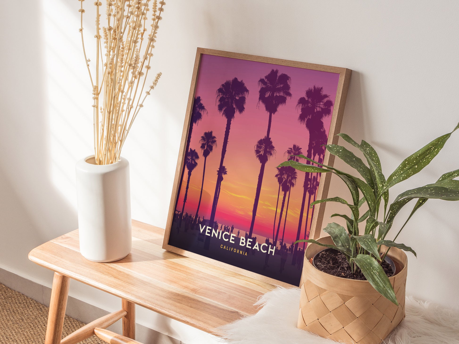 A framed Venice Beach poster with a vibrant sunset featuring palm trees, placed on a wooden bench next to a white vase with dried flowers and a potted green plant.