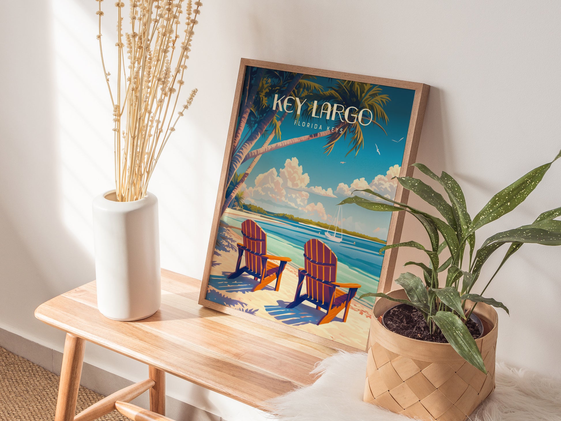 A Key Largo poster featuring a beach scene with palm trees and two chairs is placed on a wooden bench next to a potted plant and a white vase.