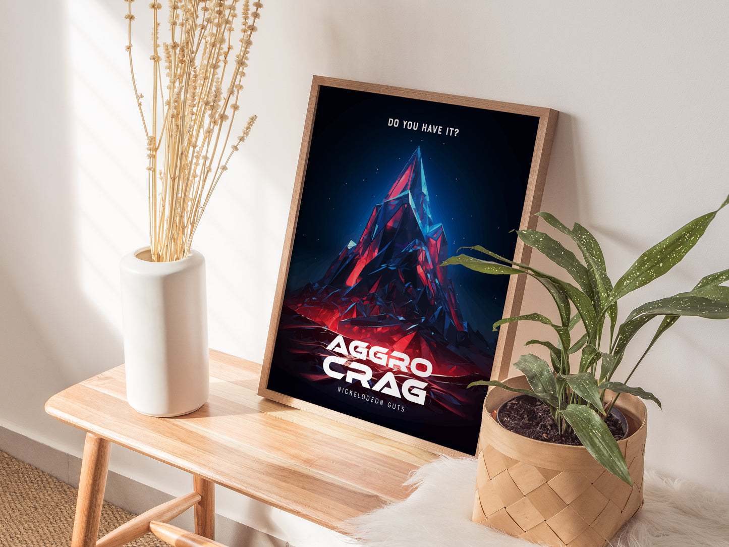 A framed Aggro Crag poster with vibrant colors leans against a wall on a wooden bench, accompanied by a white vase with dried flowers and a potted plant.