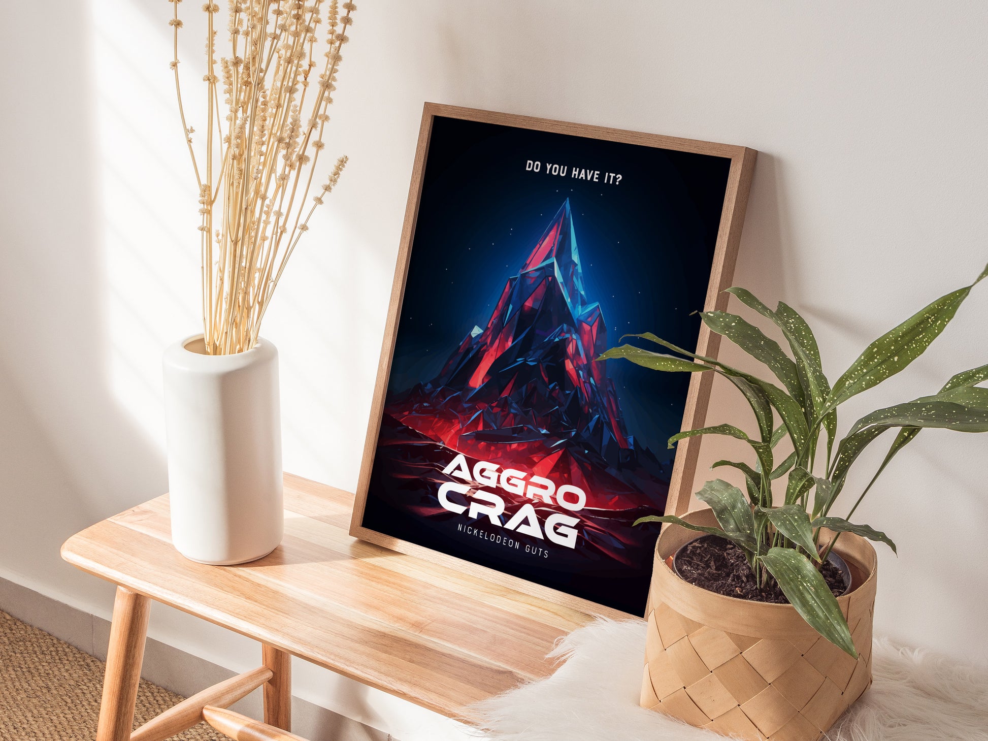A framed Aggro Crag poster with vibrant colors leans against a wall on a wooden bench, accompanied by a white vase with dried flowers and a potted plant.