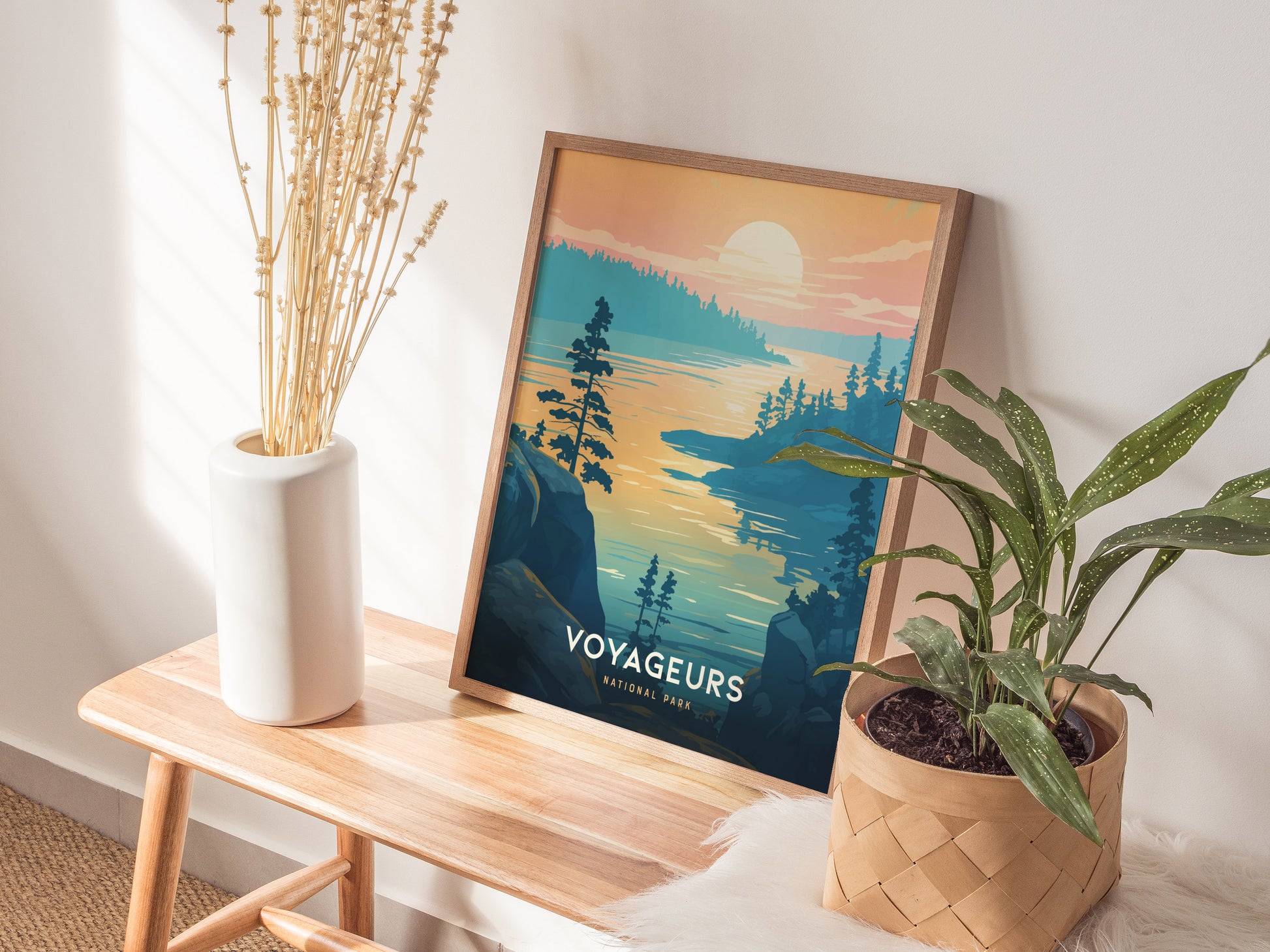 A framed Voyageurs National Park poster featuring a lake at sunset is displayed on a wooden bench, accompanied by a white vase with dried flowers and a potted green plant.