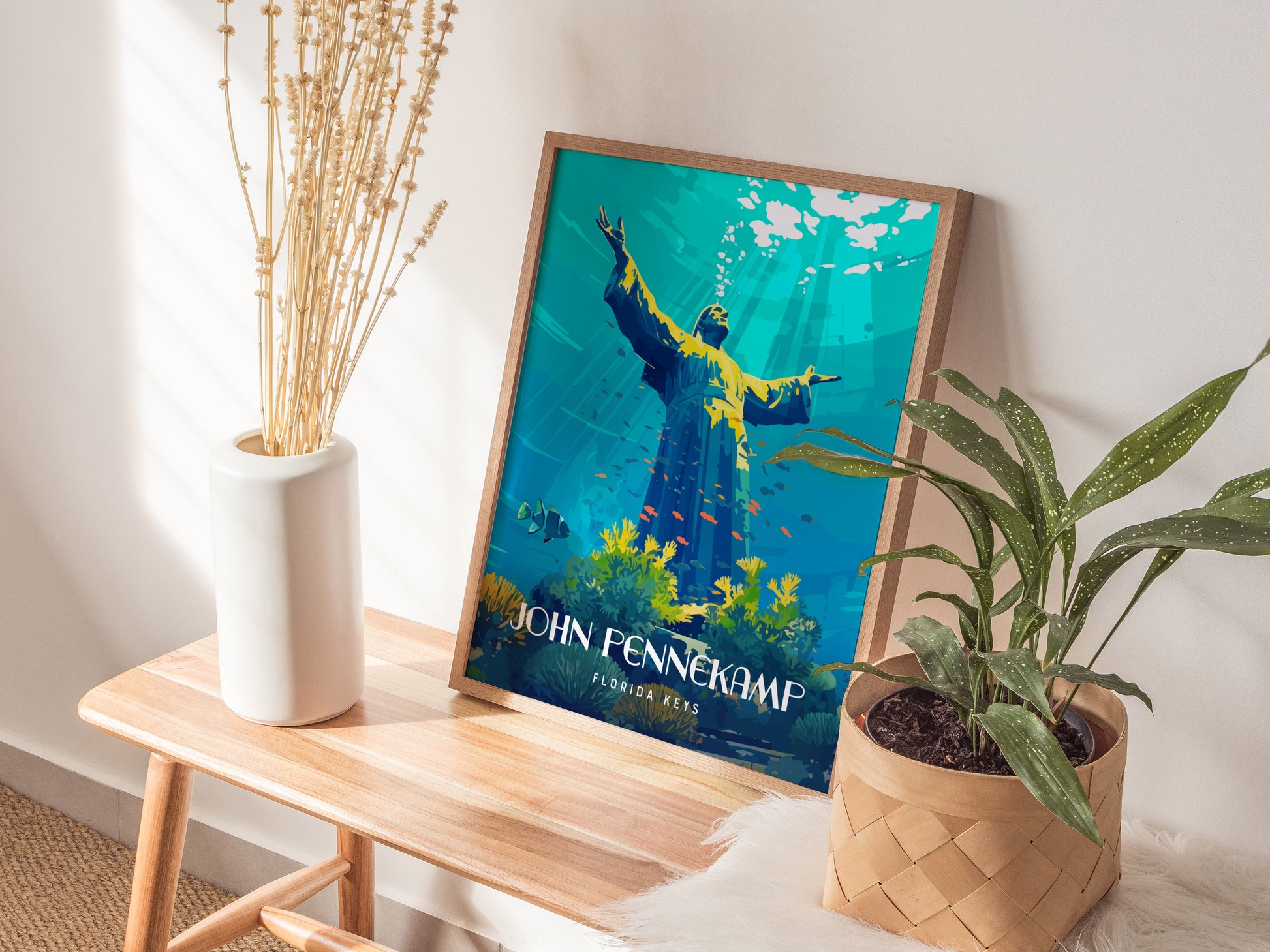 A framed John Pennekamp Coral Reef State Park poster rests on a wooden bench next to a white vase with dried flowers and a potted green plant.
