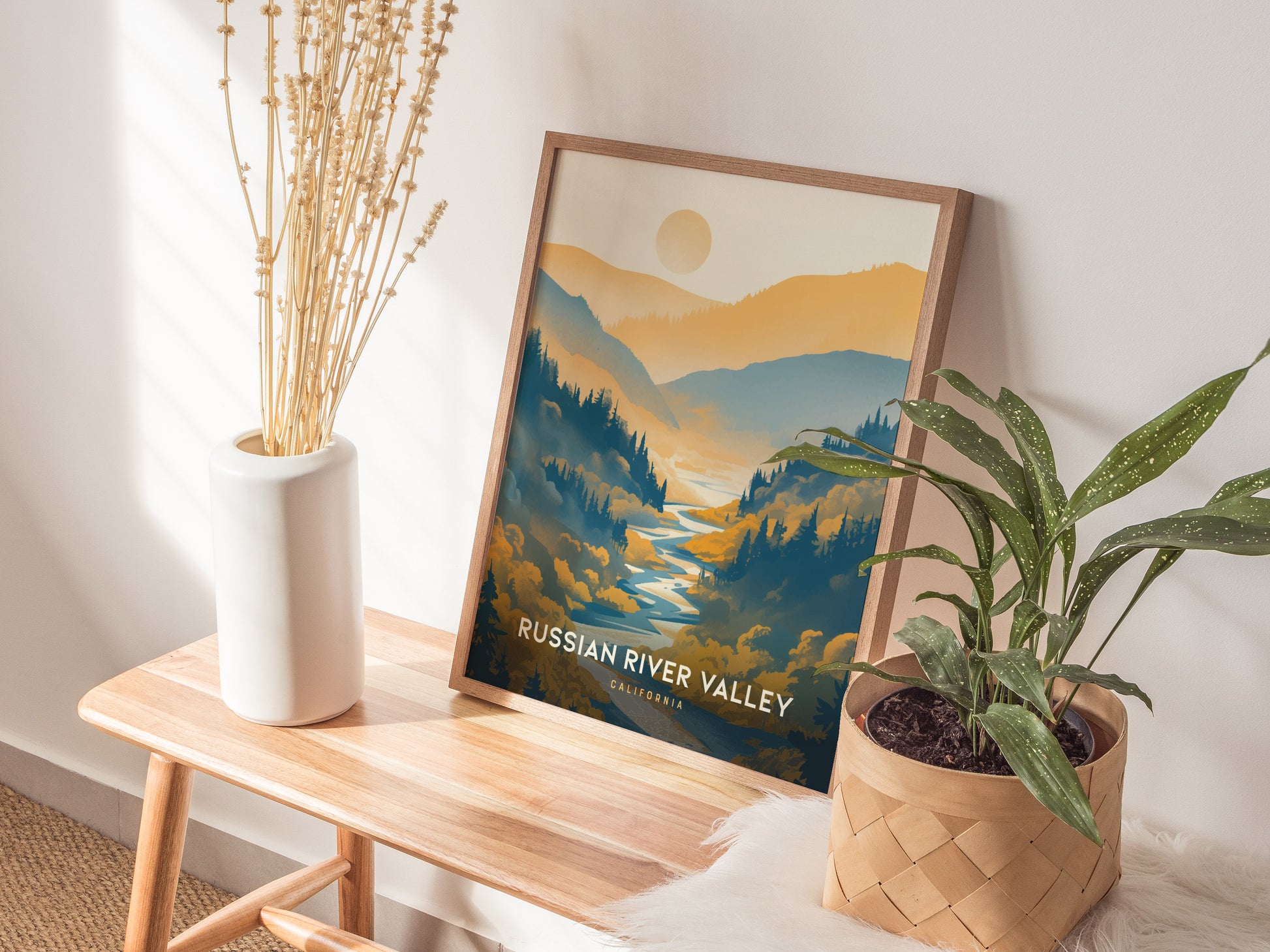 A framed poster of the Russian River Valley rests on a wooden bench, surrounded by a white vase with dried plants and a green potted plant.