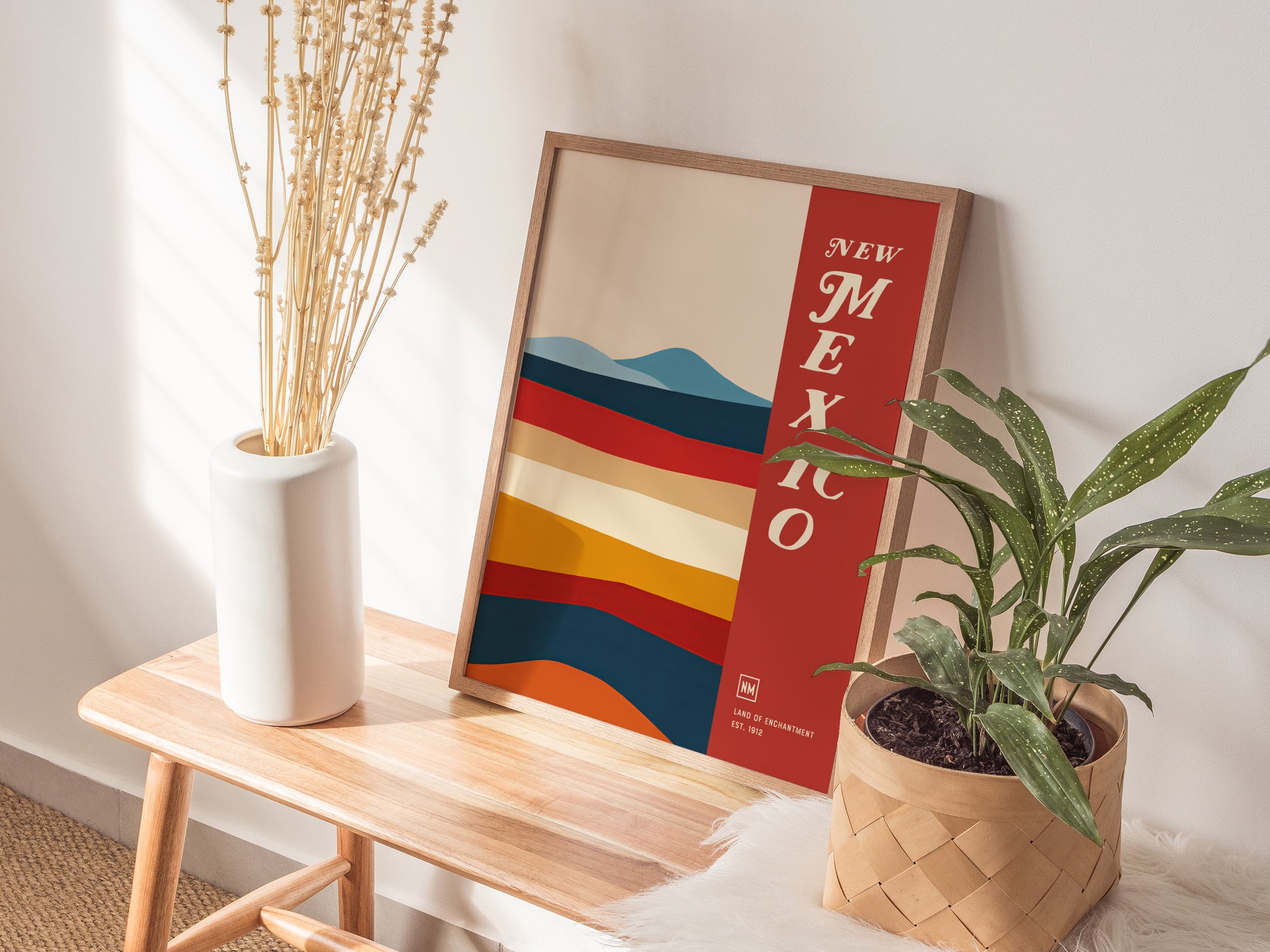 A framed New Mexico poster with colorful abstract stripes leans against a wall on a wooden bench, accompanied by a white vase with dried branches and a potted plant.