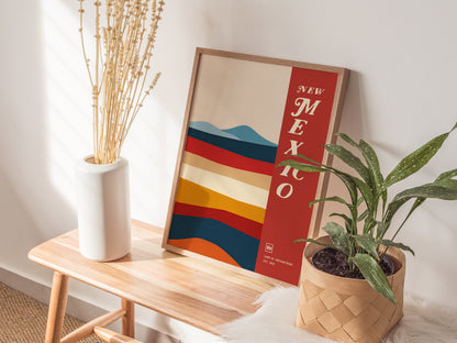 A framed New Mexico poster with colorful abstract stripes leans against a wall on a wooden bench, accompanied by a white vase with dried branches and a potted plant.
