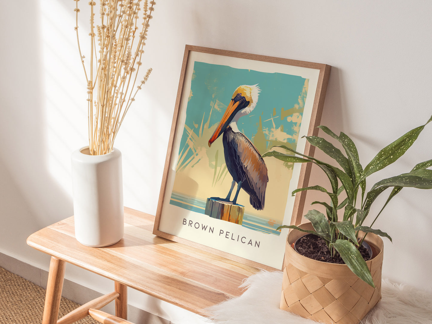 A framed Brown Pelican poster rests on a wooden bench next to a white vase with dried flowers and a potted plant, creating a cozy, natural decor setup.