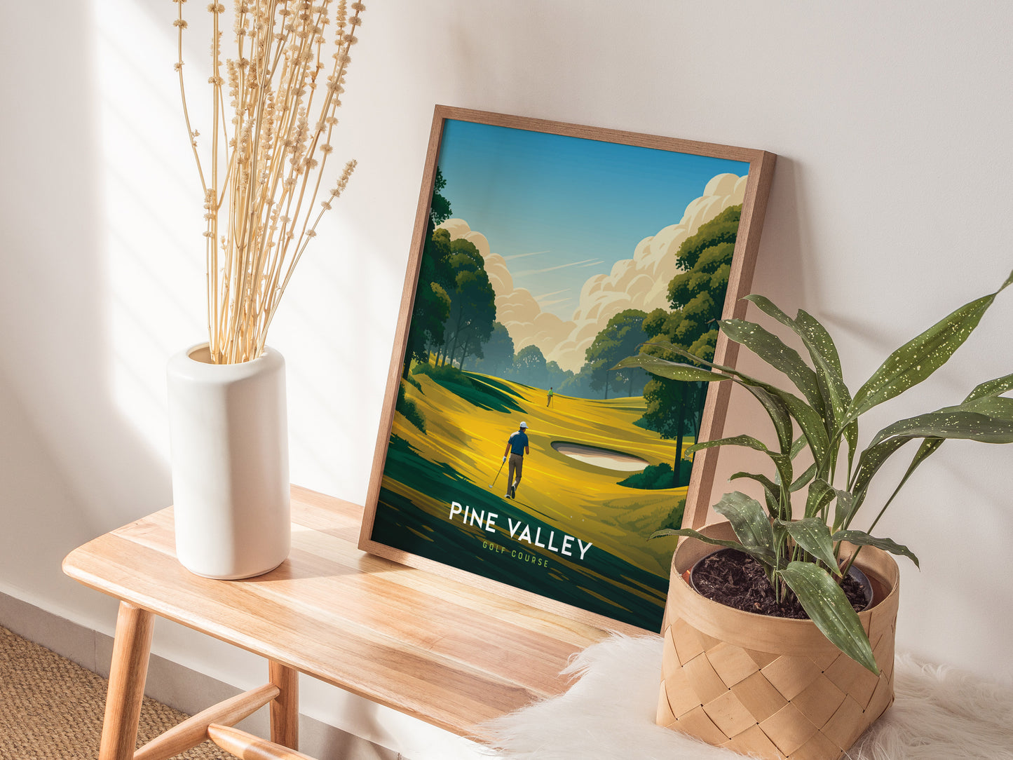 A framed Pine Valley Golf Course poster sits on a wooden bench, surrounded by a white vase with dried plants and a green potted plant, creating a cozy decor setting.