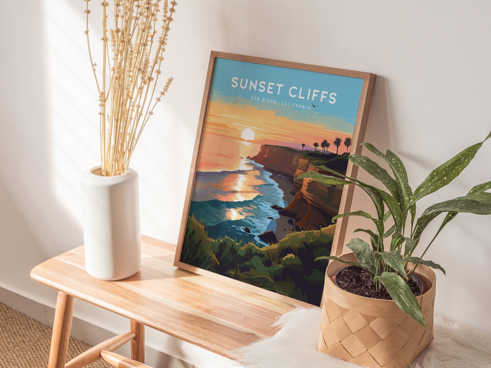 A framed poster titled Sunset Cliffs San Diego, California sits on a wooden bench, accompanied by a white vase with dried flowers and a potted plant.