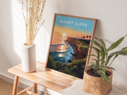 A framed poster titled Sunset Cliffs San Diego, California sits on a wooden bench, accompanied by a white vase with dried flowers and a potted plant.