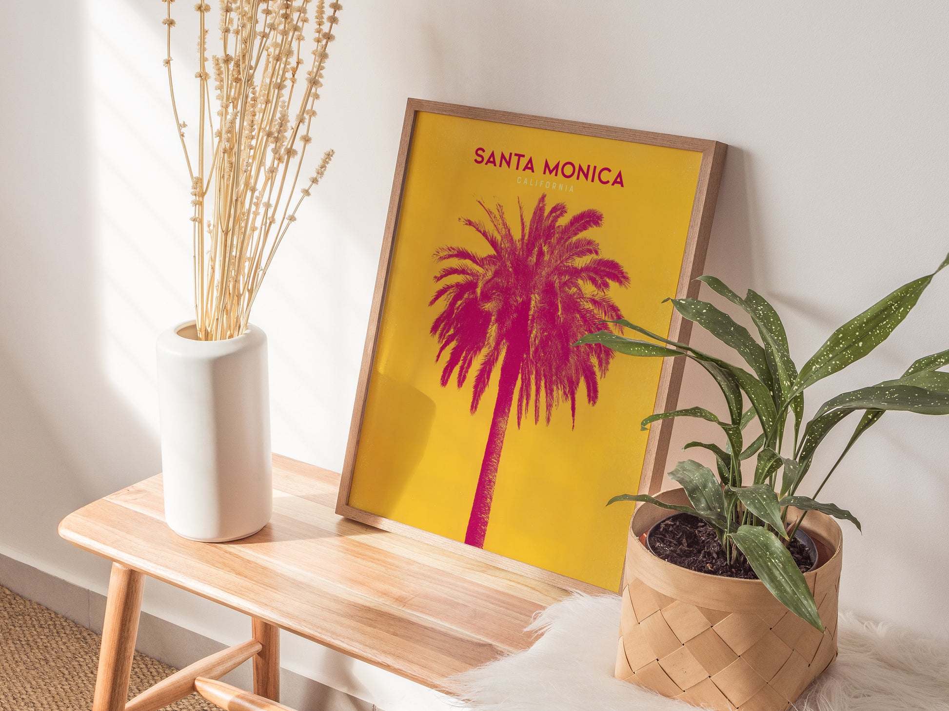 A vibrant Santa Monica poster featuring a pink palm tree on a yellow background is displayed on a wooden bench. It is surrounded by a white vase with dried stems.