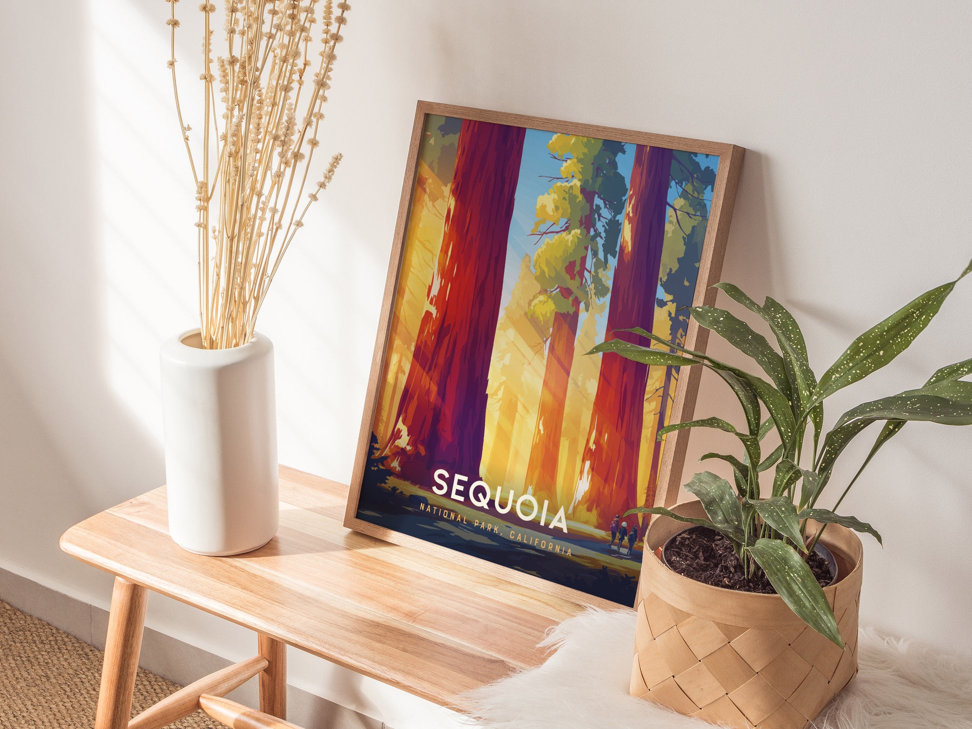 A framed Sequoia National Park poster rests on a wooden bench beside a white vase with dried flowers and a potted green plant. Sunlight casts shadows on the wall.