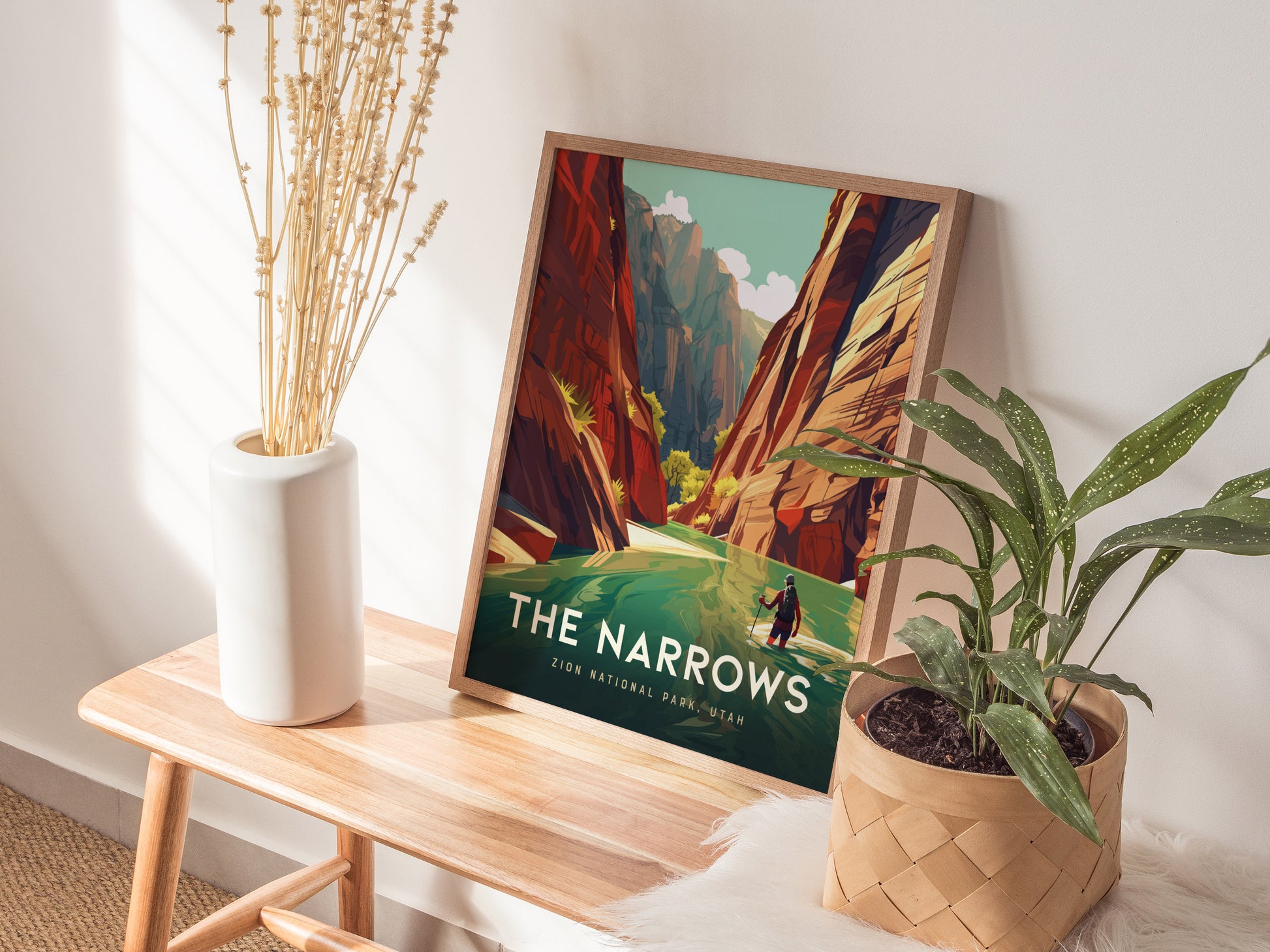 A framed poster of Zion National Parks The Narrows rests on a wooden bench, surrounded by a white vase with dried flowers and a potted green plant.