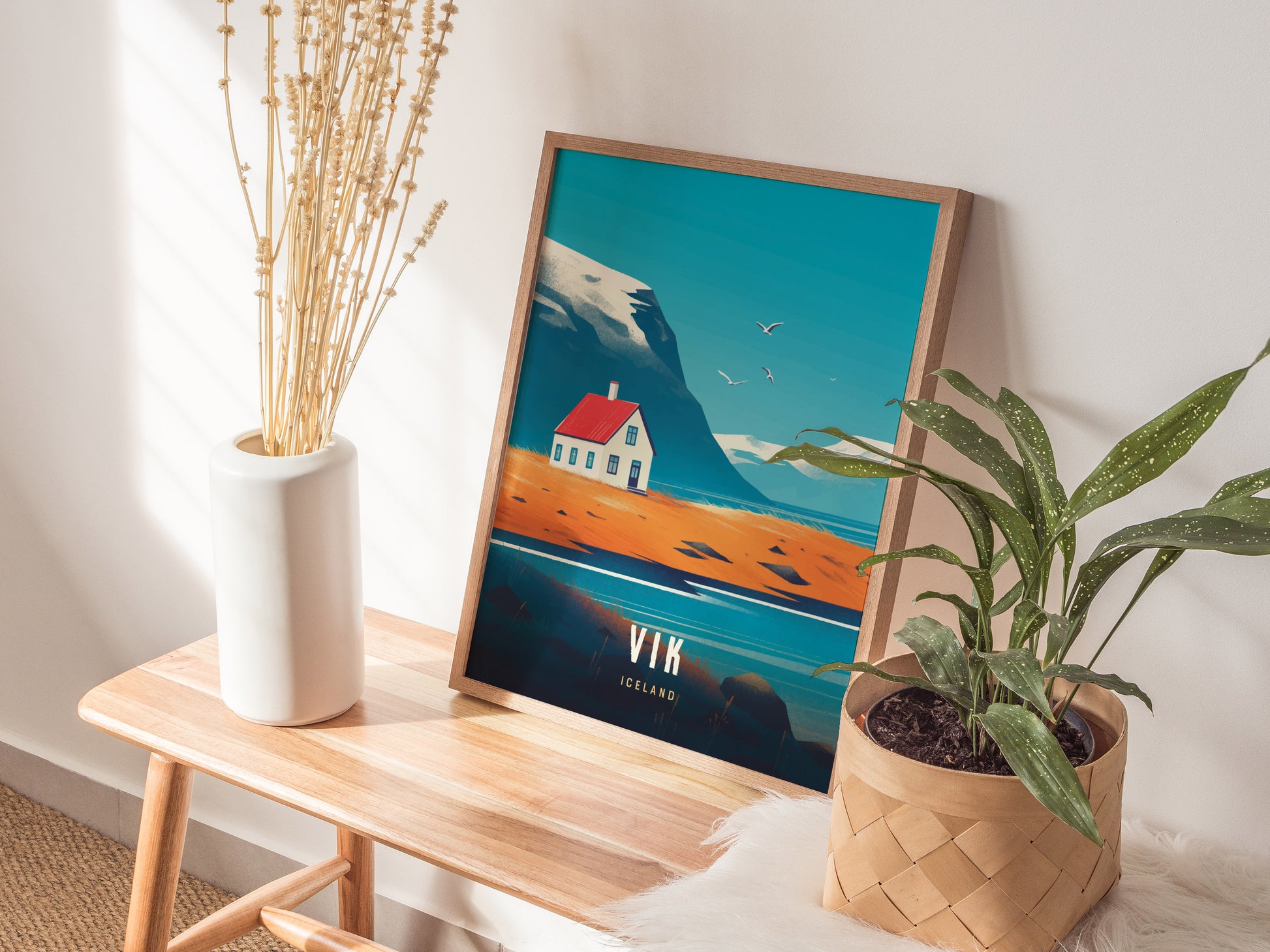 A wooden-framed poster depicting a coastal scene with a house is displayed on a bench, flanked by a white vase with dried stems and a green potted plant.