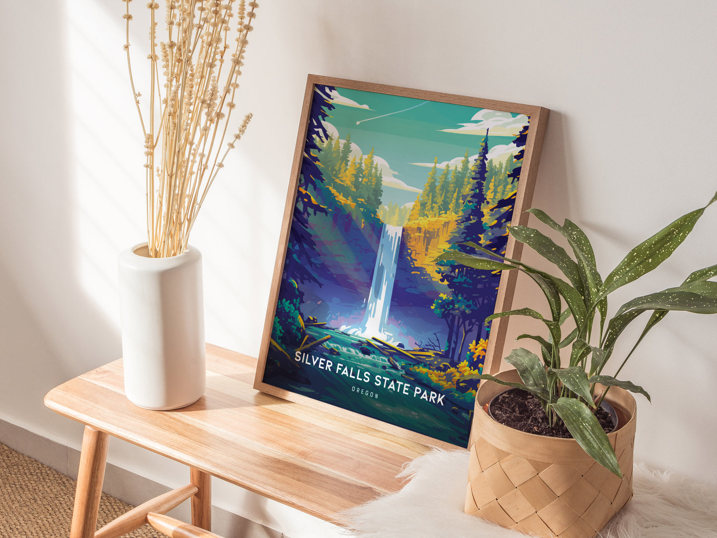 A framed Silver Falls State Park poster featuring a waterfall is placed on a wooden bench beside a white vase with dried flowers and a potted green plant.