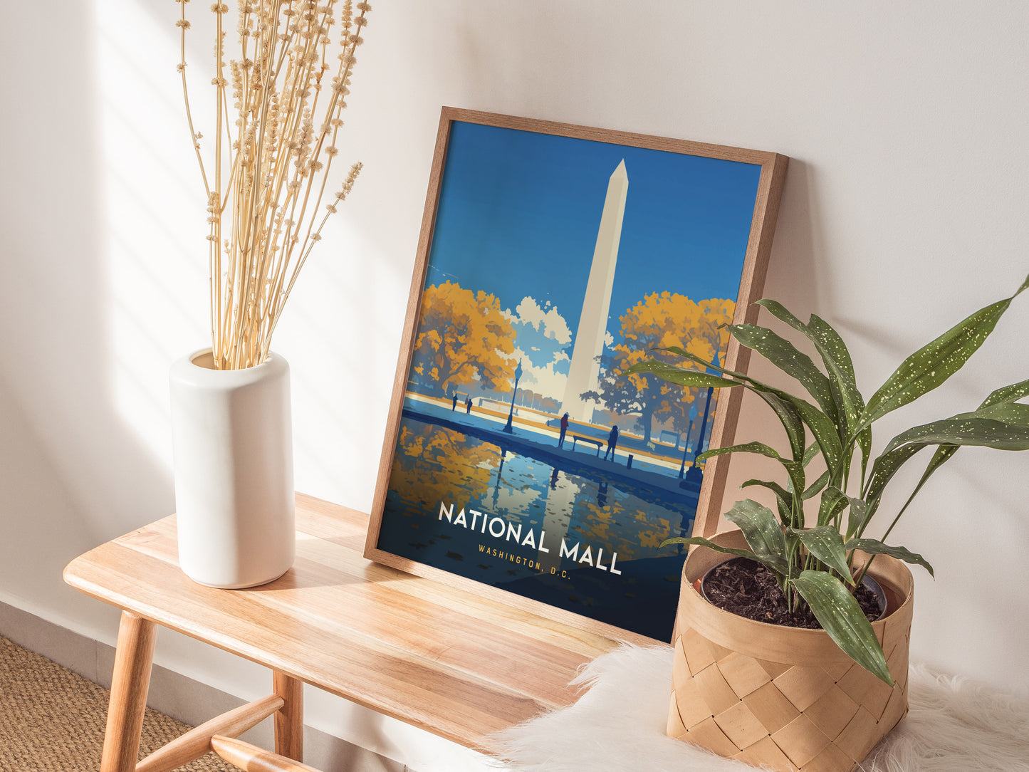 A framed National Mall poster featuring the Washington Monument is placed on a wooden bench beside a white vase with dried stems and a potted plant.