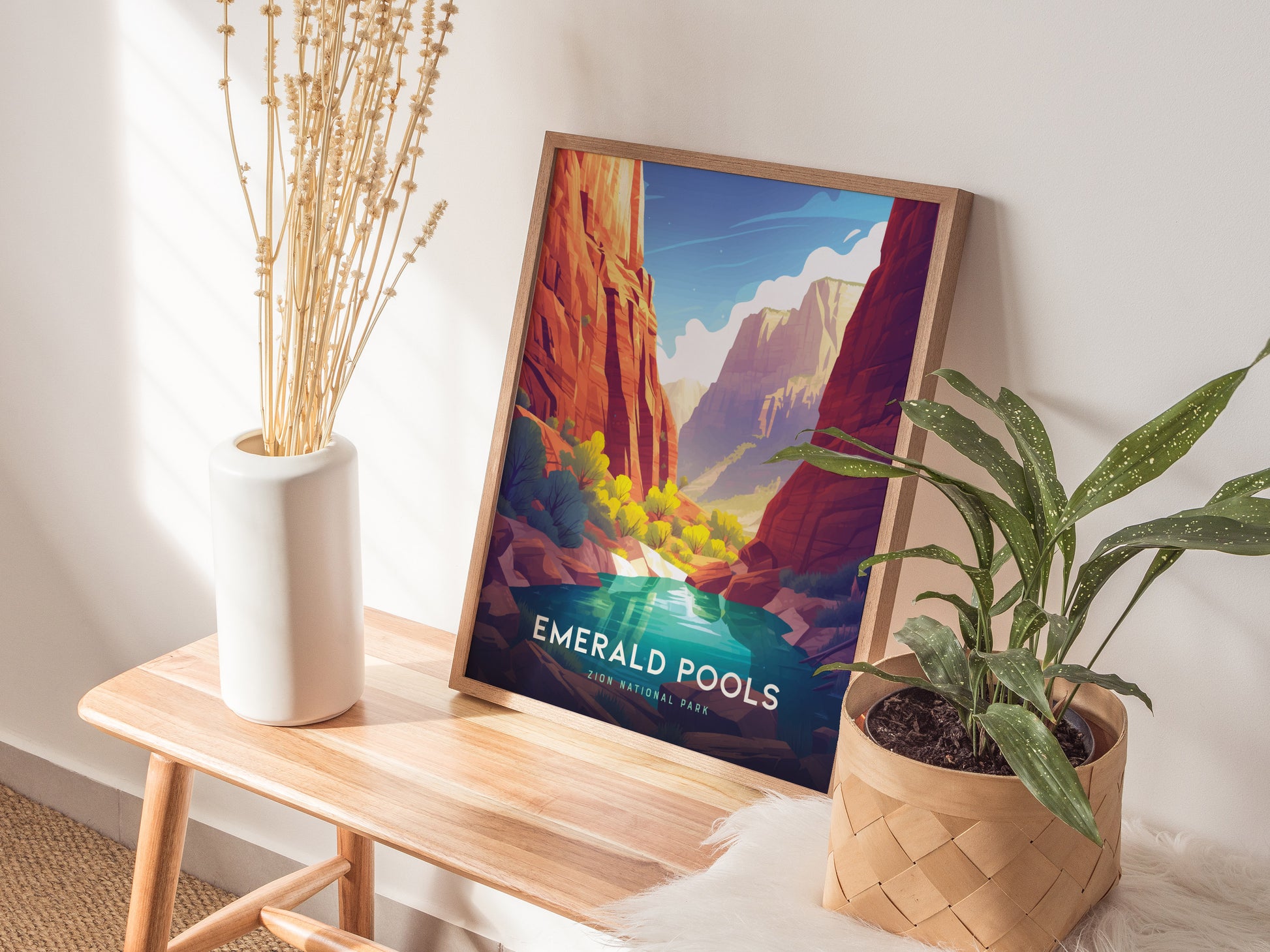 A framed poster of Emerald Pools in Zion National Park rests on a wooden bench, accompanied by a white vase with dried stems and a potted green plant.