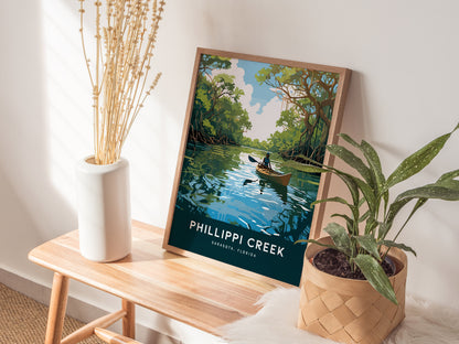 A framed poster of a kayaker on a serene creek leans against a wall, alongside a vase with dried flowers and a potted plant on a wooden bench.