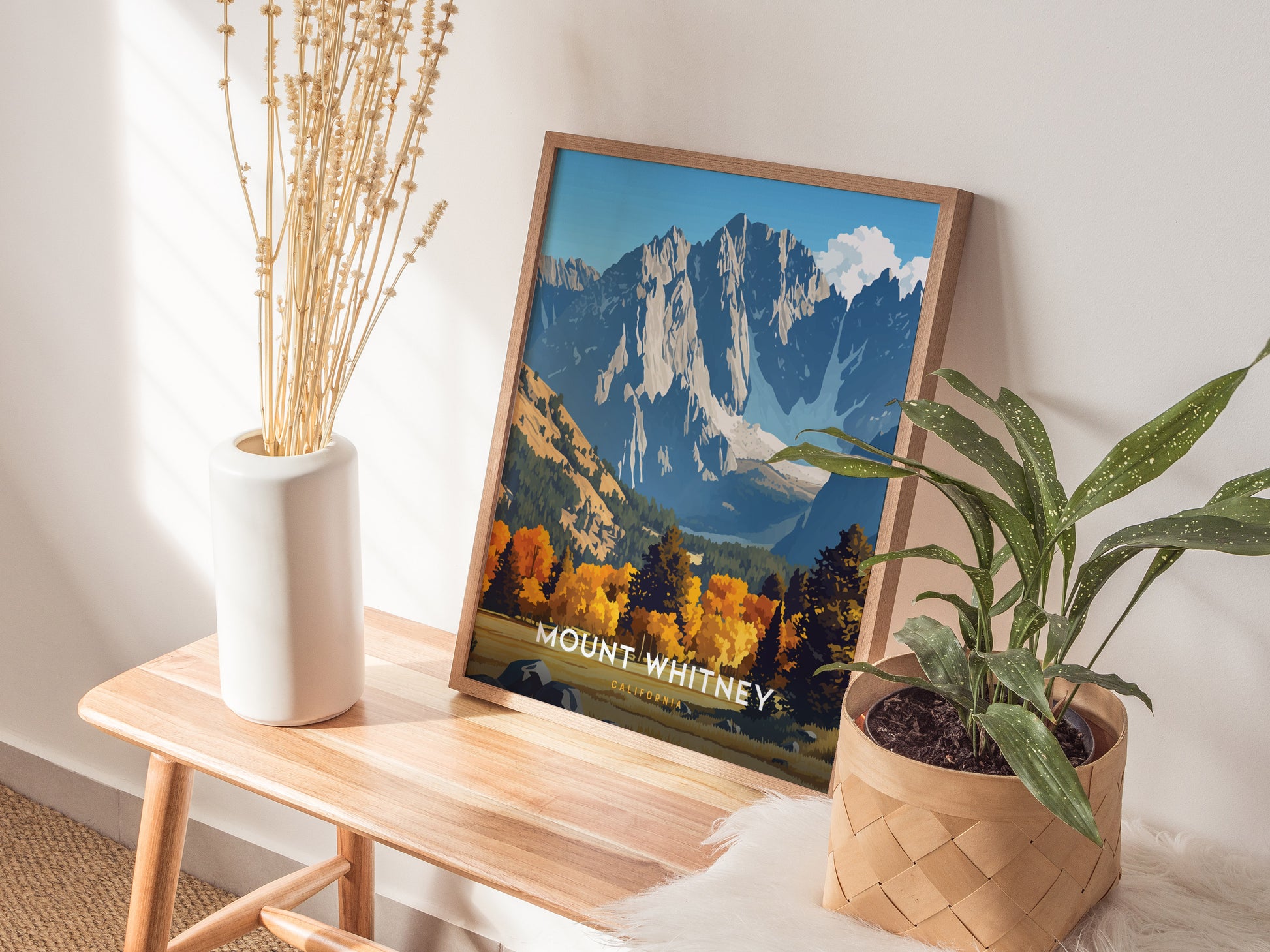 A framed Mount Whitney poster rests on a wooden bench, accompanied by a white vase with dried flowers and a potted green plant, creating a cozy, natural decor setting.