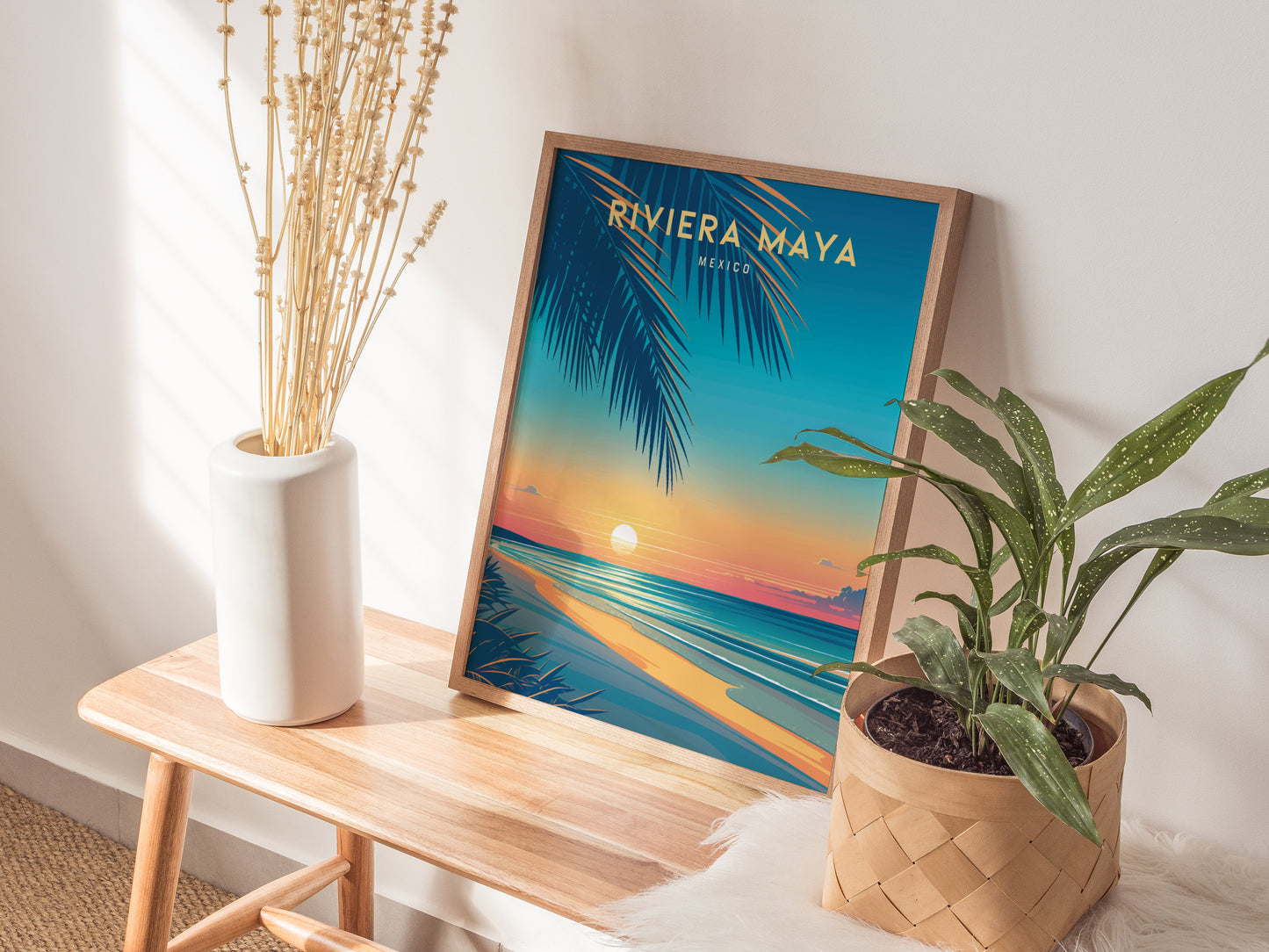 A colorful Riviera Maya travel poster is displayed on a wooden bench, surrounded by a tall white vase with dried branches and a potted green plant.
