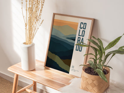 A framed abstract poster of Colorados mountains rests on a wooden bench, accompanied by a white vase with dried stems and a potted green plant.