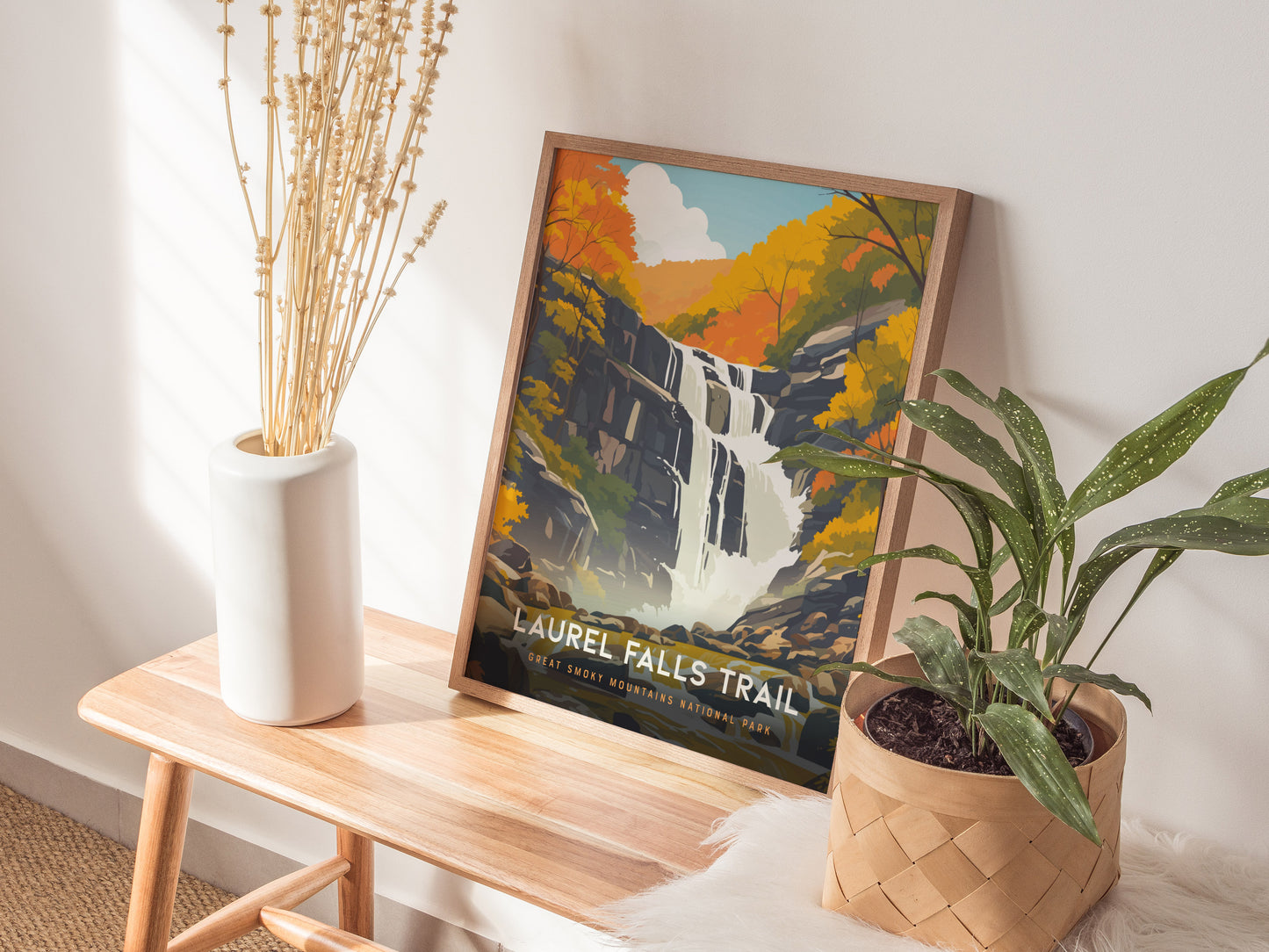 A framed Laurel Falls Trail poster sits on a wooden bench, surrounded by a white vase with dried flowers and a potted plant, in a sunlit room.