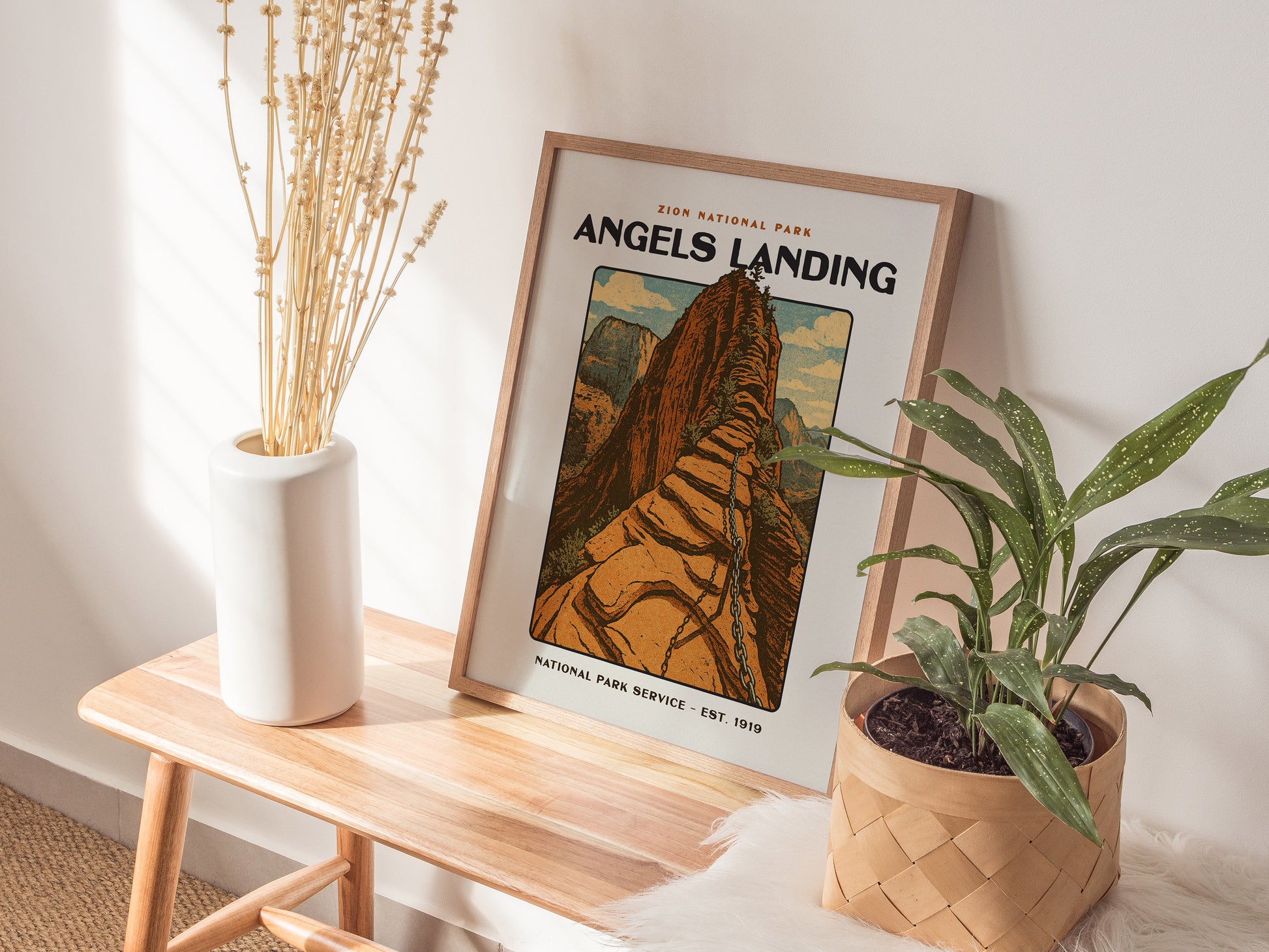 A framed vintage-style Angels Landing poster leans against a wall on a wooden bench, accompanied by a white vase with dried flowers and a potted green plant.