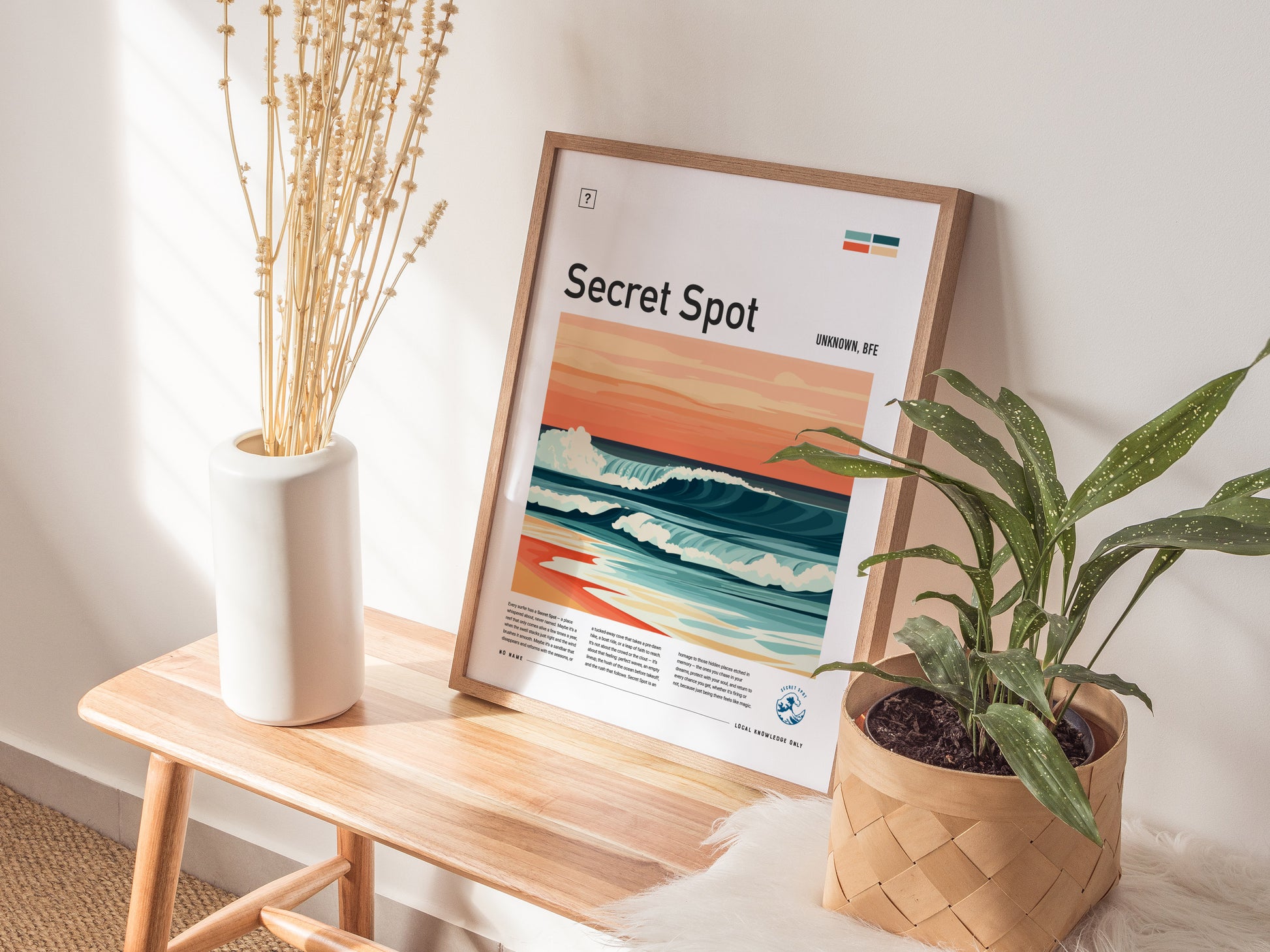 A framed surf-themed poster titled Secret Spot rests on a wooden bench beside a white vase with dried flowers and a potted plant. Soft sunlight illuminates the scene.
