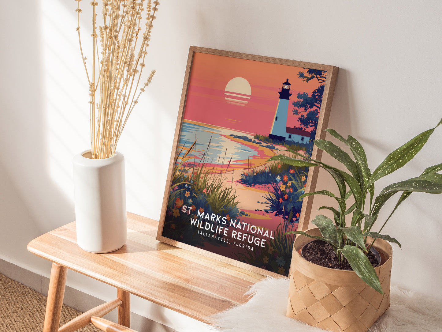 A framed poster of St. Marks Wildlife Refuge sits on a wooden table beside a white vase with dried flowers and a potted plant, capturing a lighthouse scene at sunset.