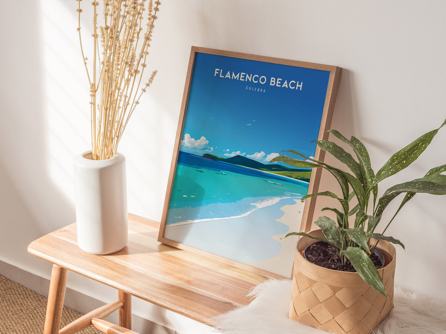 A framed Flamenco Beach poster from Culebra, Puerto Rico, rests on a wooden bench beside a tall white vase with dried flowers and a leafy green potted plant.