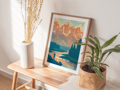 A framed Grand Teton poster leans on a wall beside a white vase with dried flowers and a potted green plant, all placed on a wooden bench.