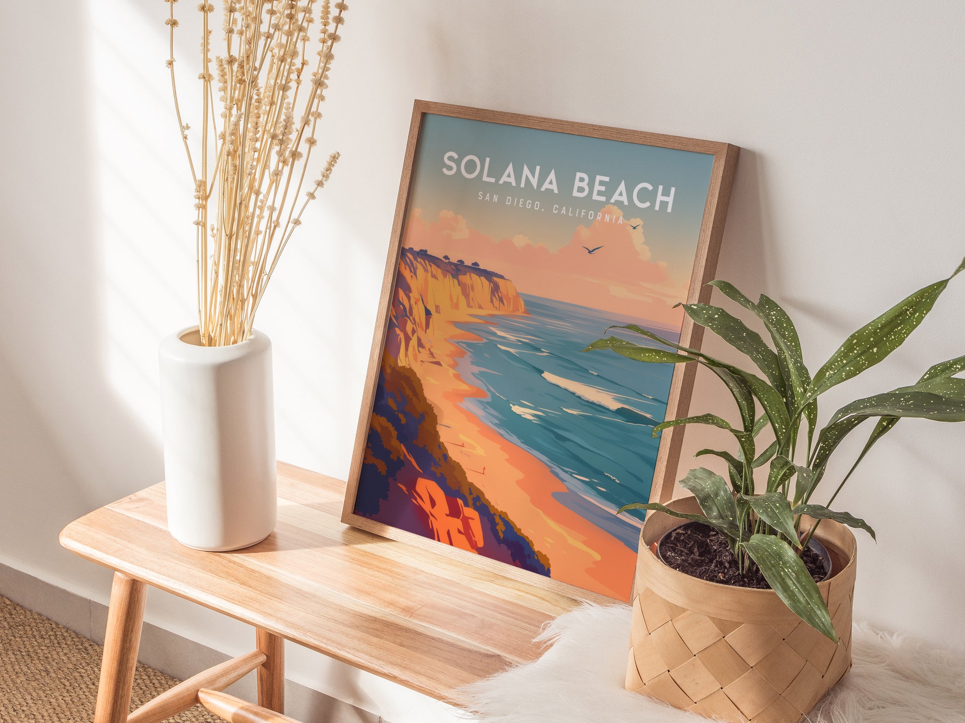 A framed Solana Beach poster depicting coastal bluffs and ocean waves is displayed on a wooden bench, accompanied by a white vase with dried stems and a potted plant.