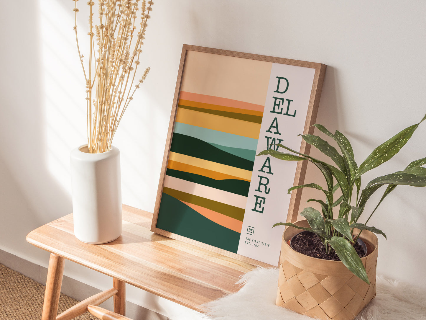 A minimalist Delaware poster with abstract, earthy tones leans against a white wall on a wooden bench, accompanied by a white vase with dried flowers and a potted plant.