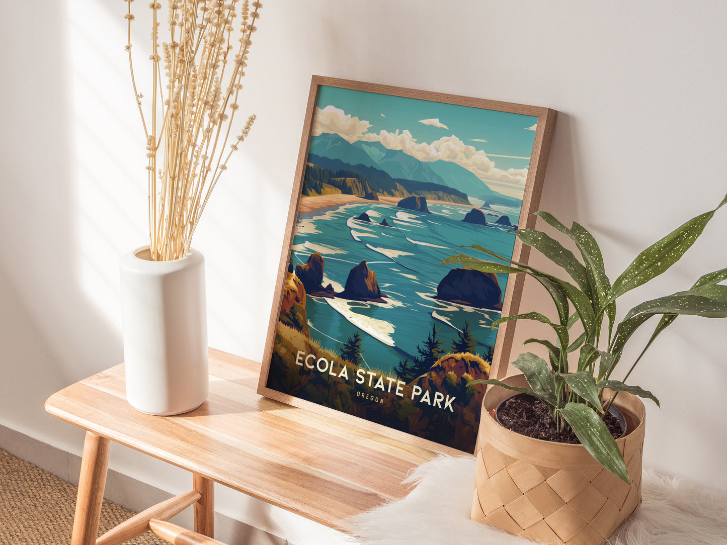 A framed Ecola State Park poster sits on a wooden bench, accompanied by a white vase with dried stems and a potted green plant.