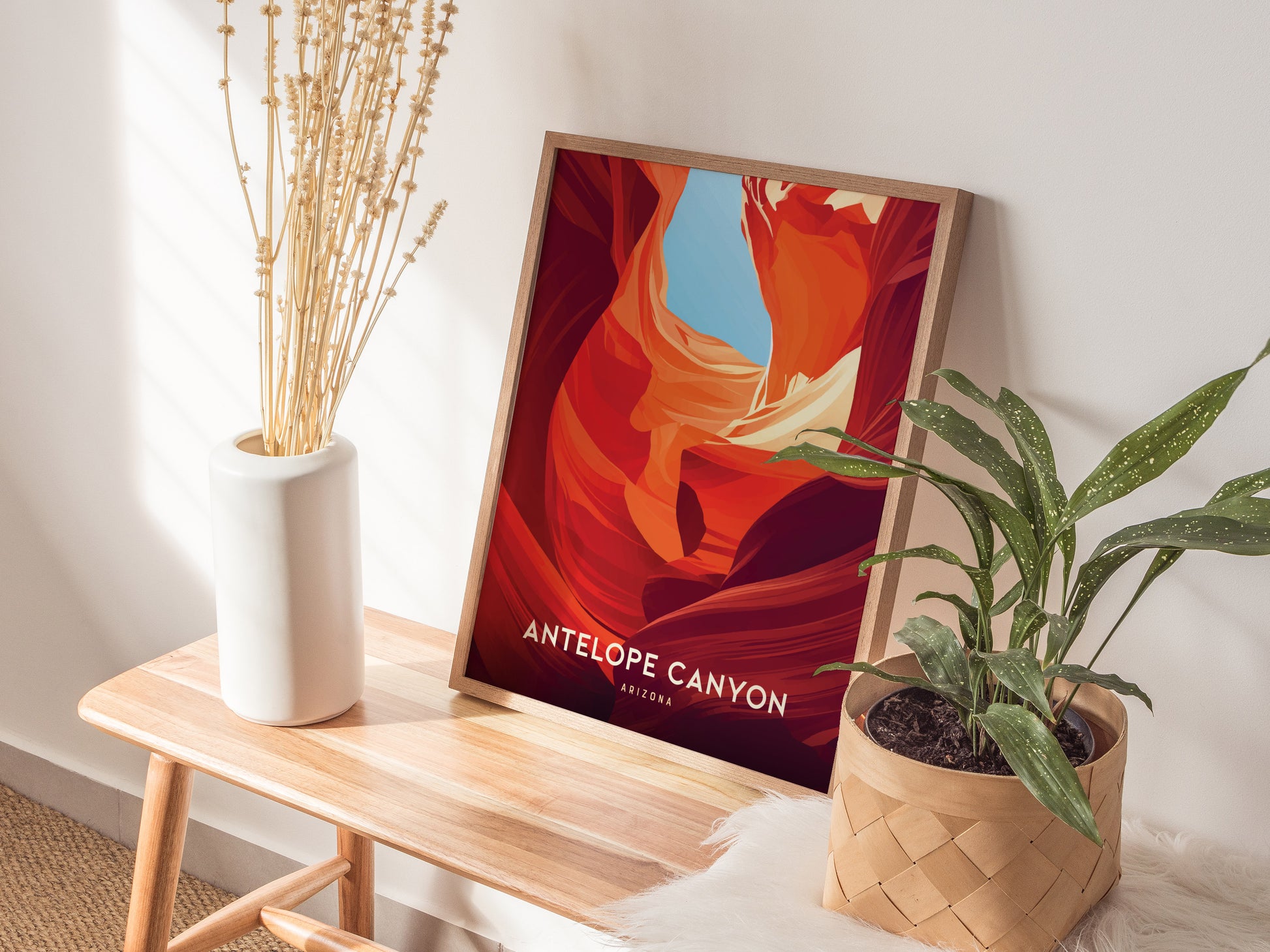 A framed Antelope Canyon poster rests on a wooden bench beside a potted green plant and a white vase with dried flowers. Sunlight casts soft shadows on the wall.