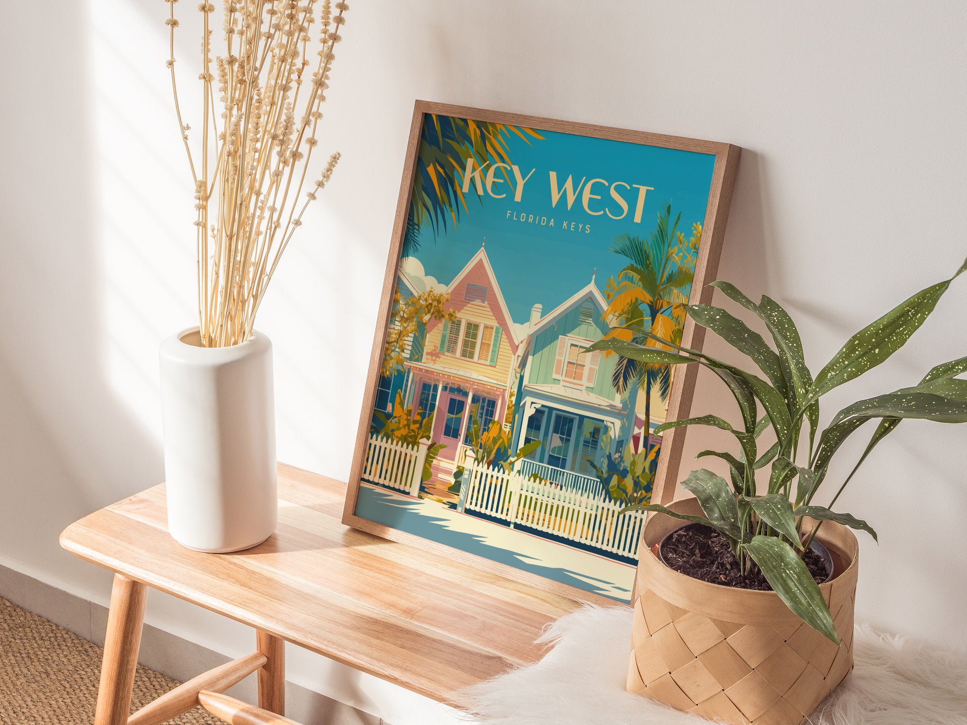 A framed Key West poster featuring pastel houses and palm trees sits on a wooden bench, complemented by a white vase with dried flowers and a potted green plant.