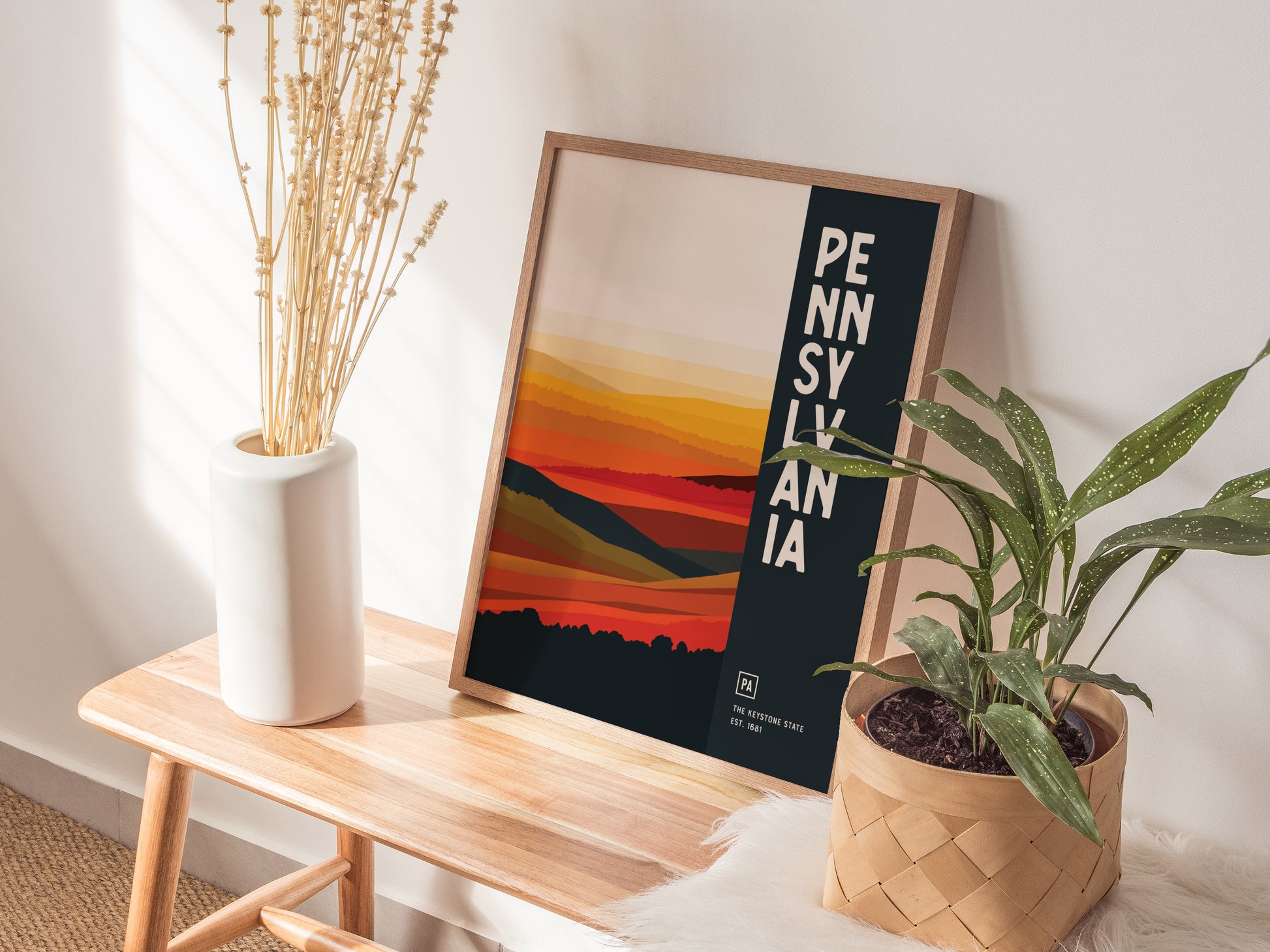 A Pennsylvania Keystone State poster with abstract landscape art is displayed on a wooden bench, accompanied by a white vase with dried flowers and a potted plant.