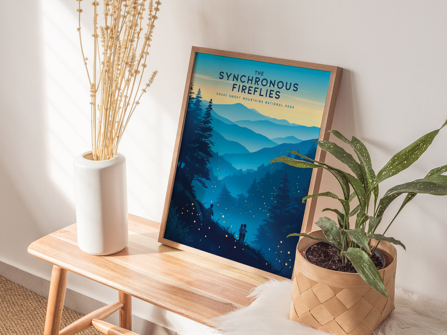 A framed poster titled The Synchronous Fireflies sits on a wooden bench, depicting a scenic view of the Great Smoky Mountains at dusk. Beside it, a white vase with dried flowers and a potted plant add a natural touch.