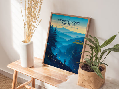 A framed poster titled The Synchronous Fireflies sits on a wooden bench, depicting a scenic view of the Great Smoky Mountains at dusk. Beside it, a white vase with dried flowers and a potted plant add a natural touch.