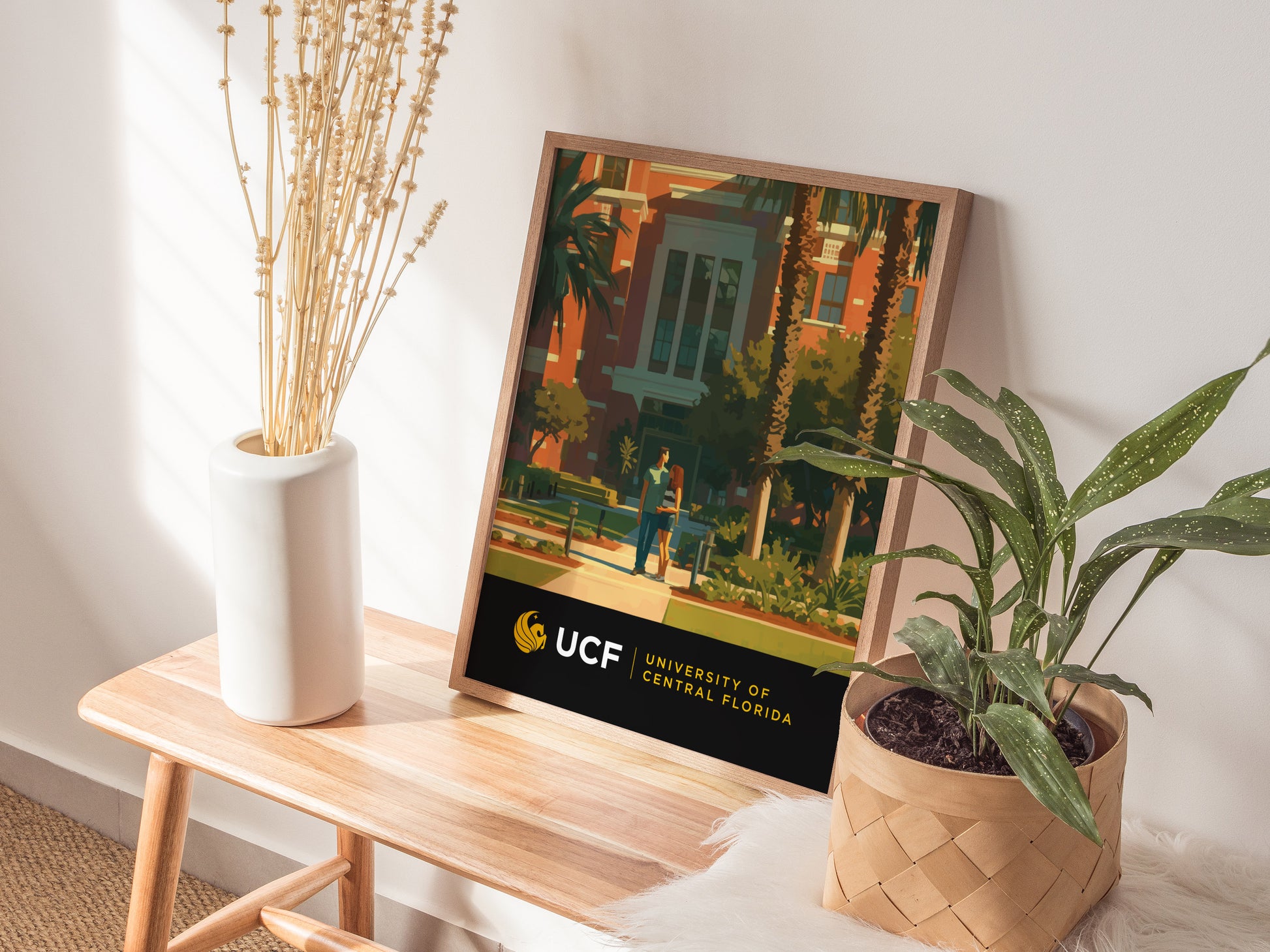 A University of Central Florida poster is displayed on a wooden bench beside a white vase with dried flowers and a potted green plant, creating a cozy, decorative setting.