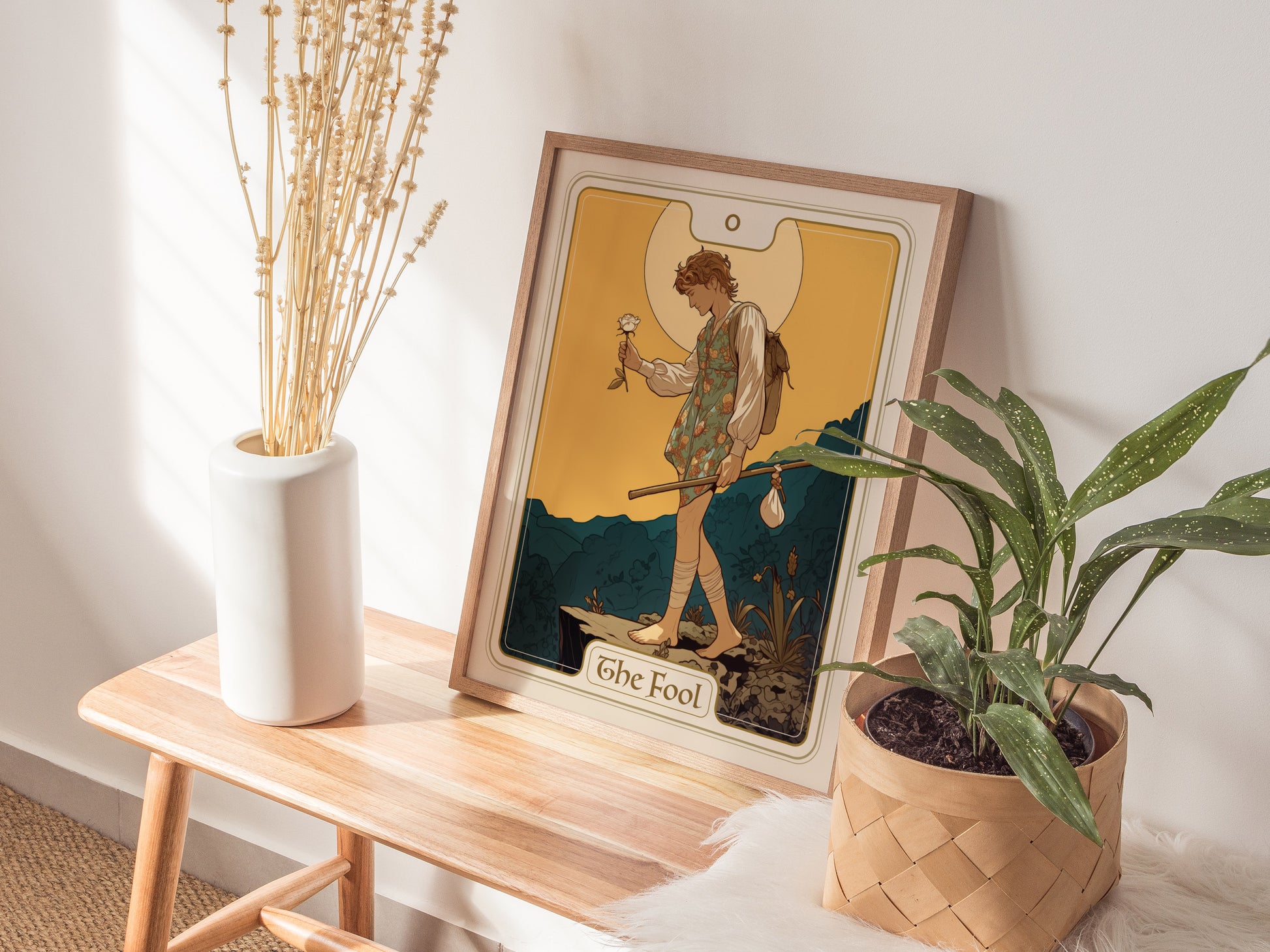 A framed Fool Tarot Card poster rests on a wooden bench beside a white vase with dried flowers and a potted plant, creating a serene and artistic display.
