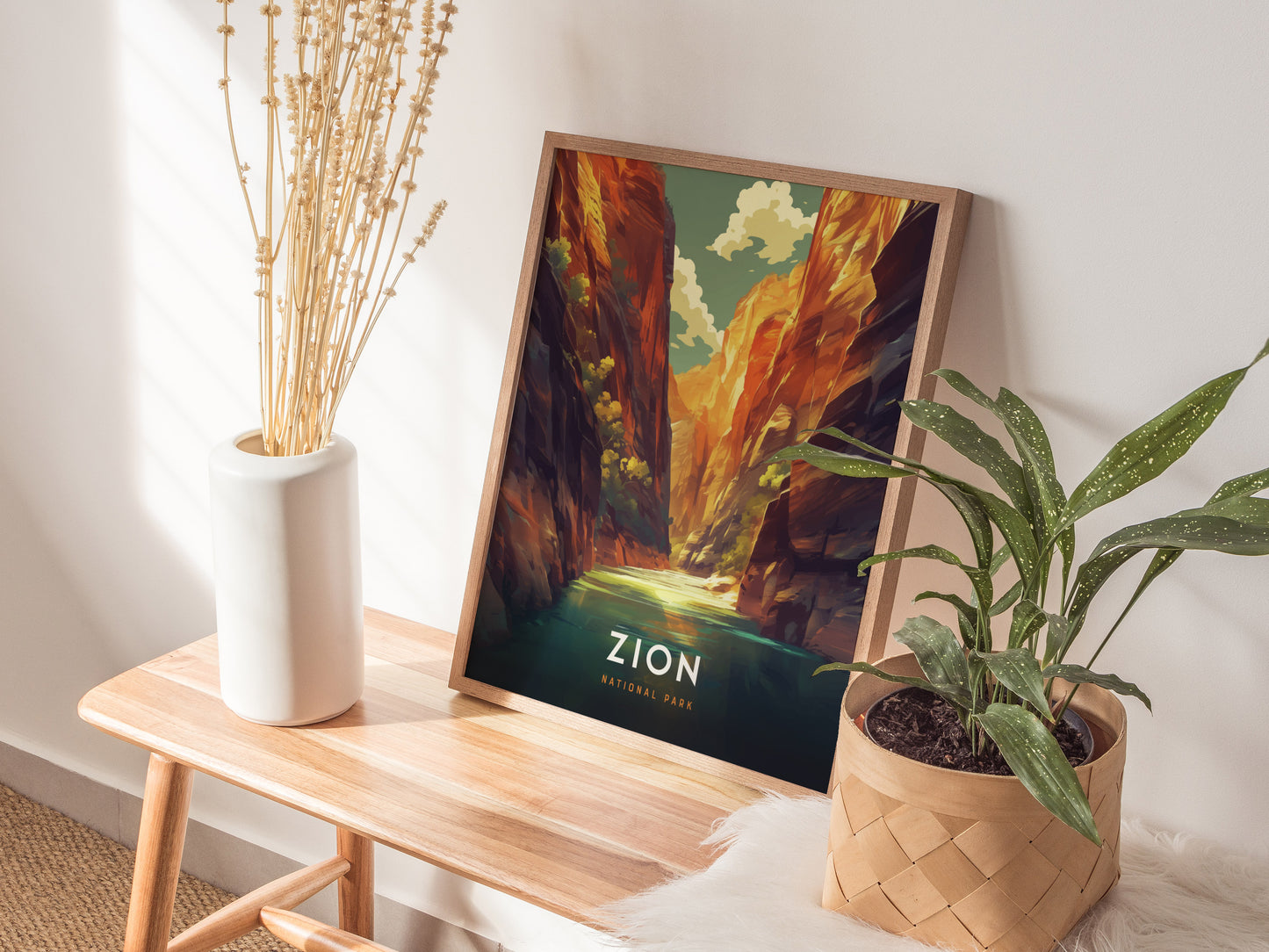A framed Zion National Park poster depicting The Narrows rests on a wooden table, accompanied by a vase of dried flowers and a potted plant. Sunlight filters through nearby.