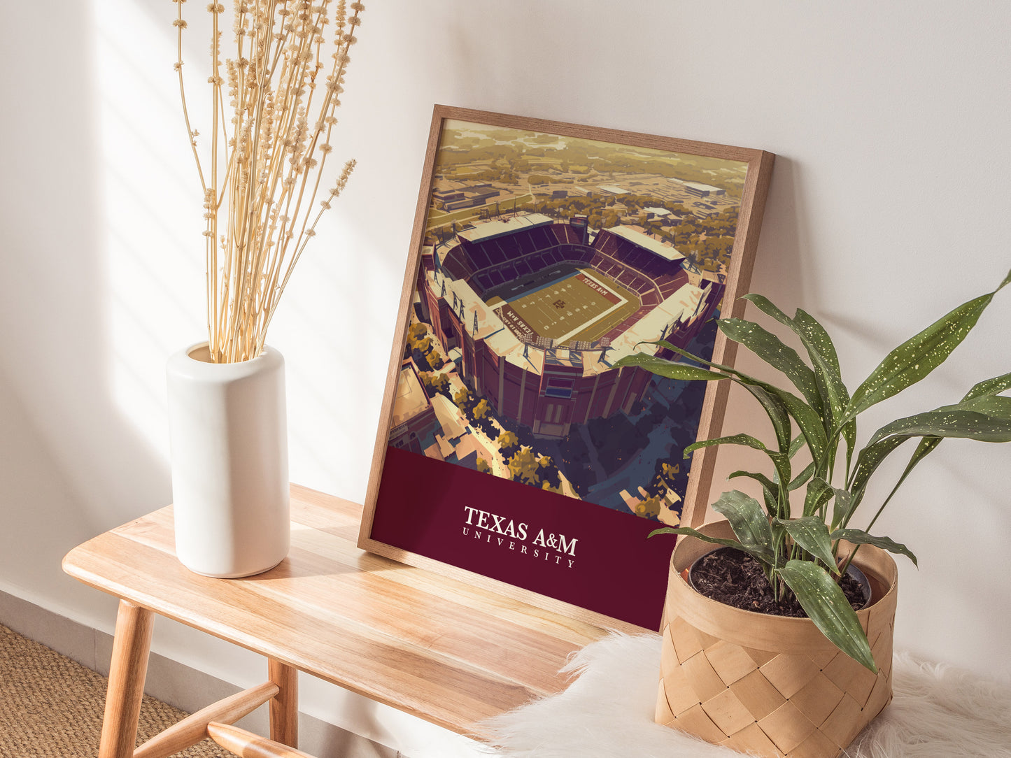 A framed Kyle Field poster depicting Texas A&M University stadium leans against a wall on a wooden table with a vase of dried flowers and a potted plant nearby.