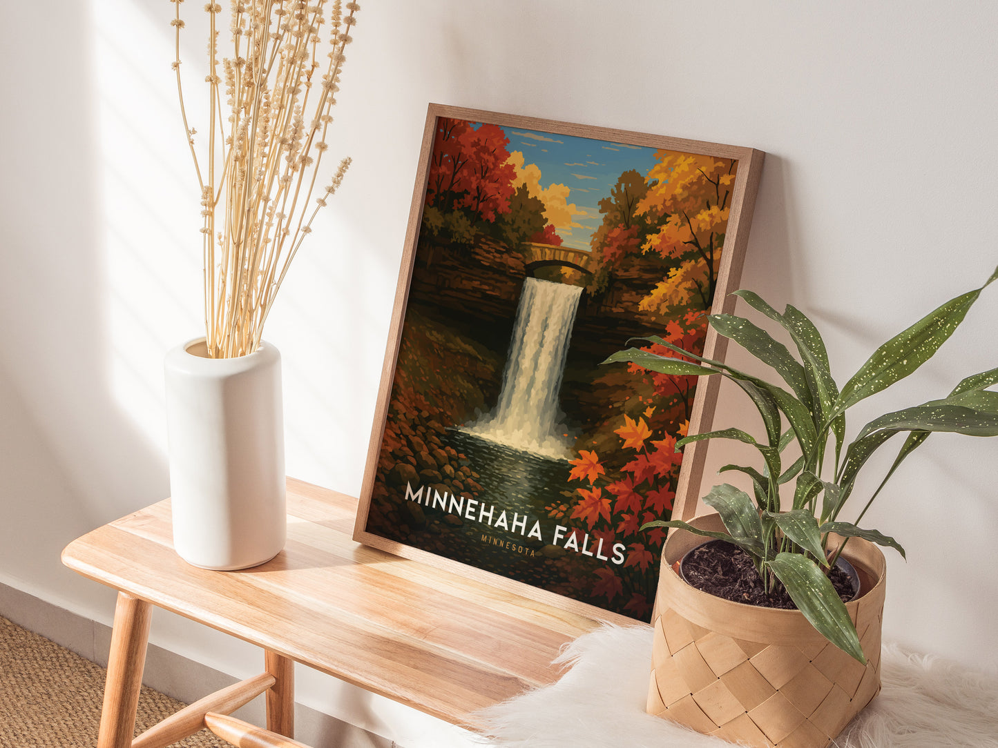 A framed Minnehaha Falls poster depicting a waterfall surrounded by autumn foliage is displayed on a wooden table, accompanied by a white vase with dried flowers and a potted plant.