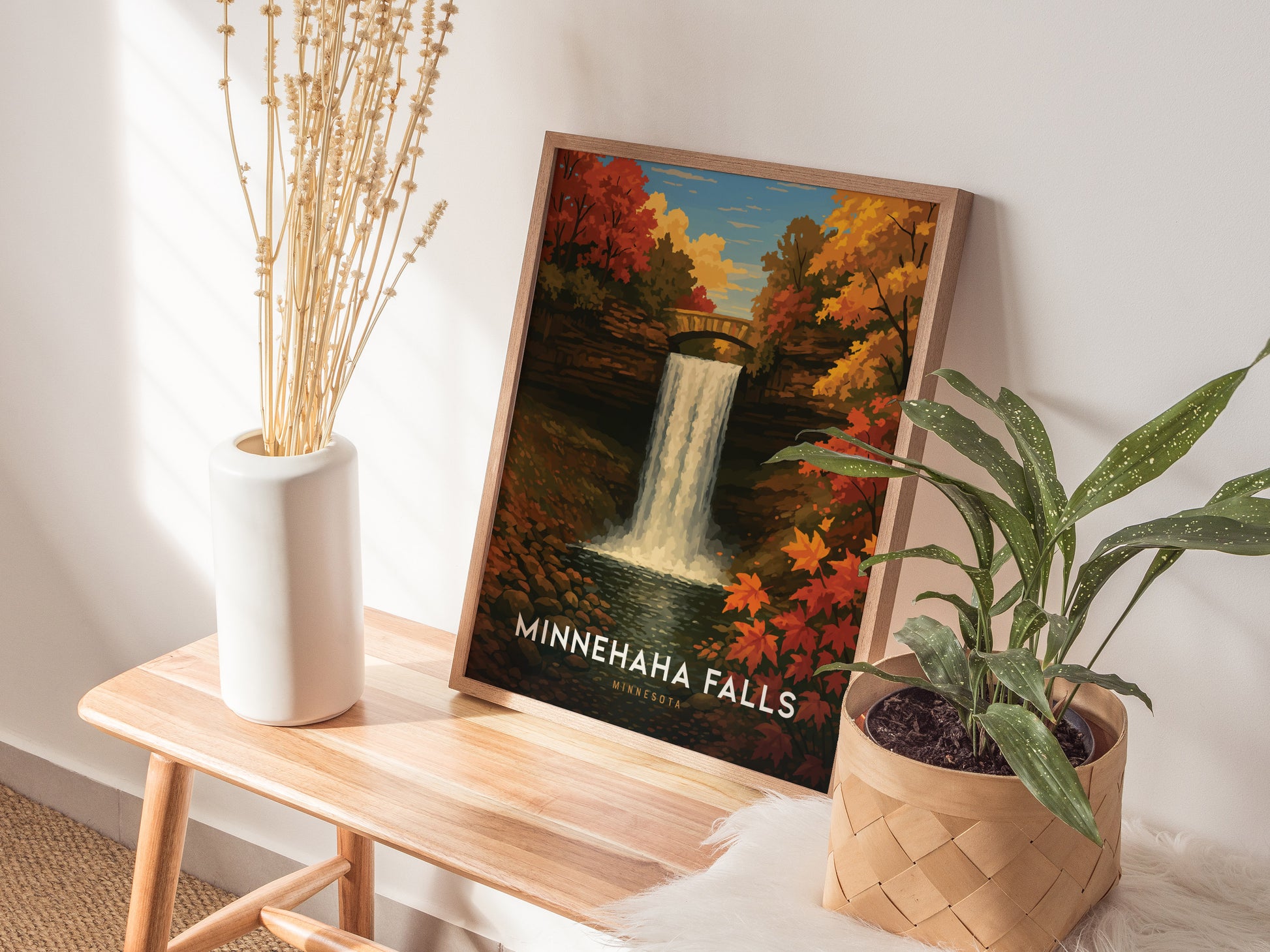 A framed Minnehaha Falls poster depicting a waterfall surrounded by autumn foliage is displayed on a wooden table, accompanied by a white vase with dried flowers and a potted plant.