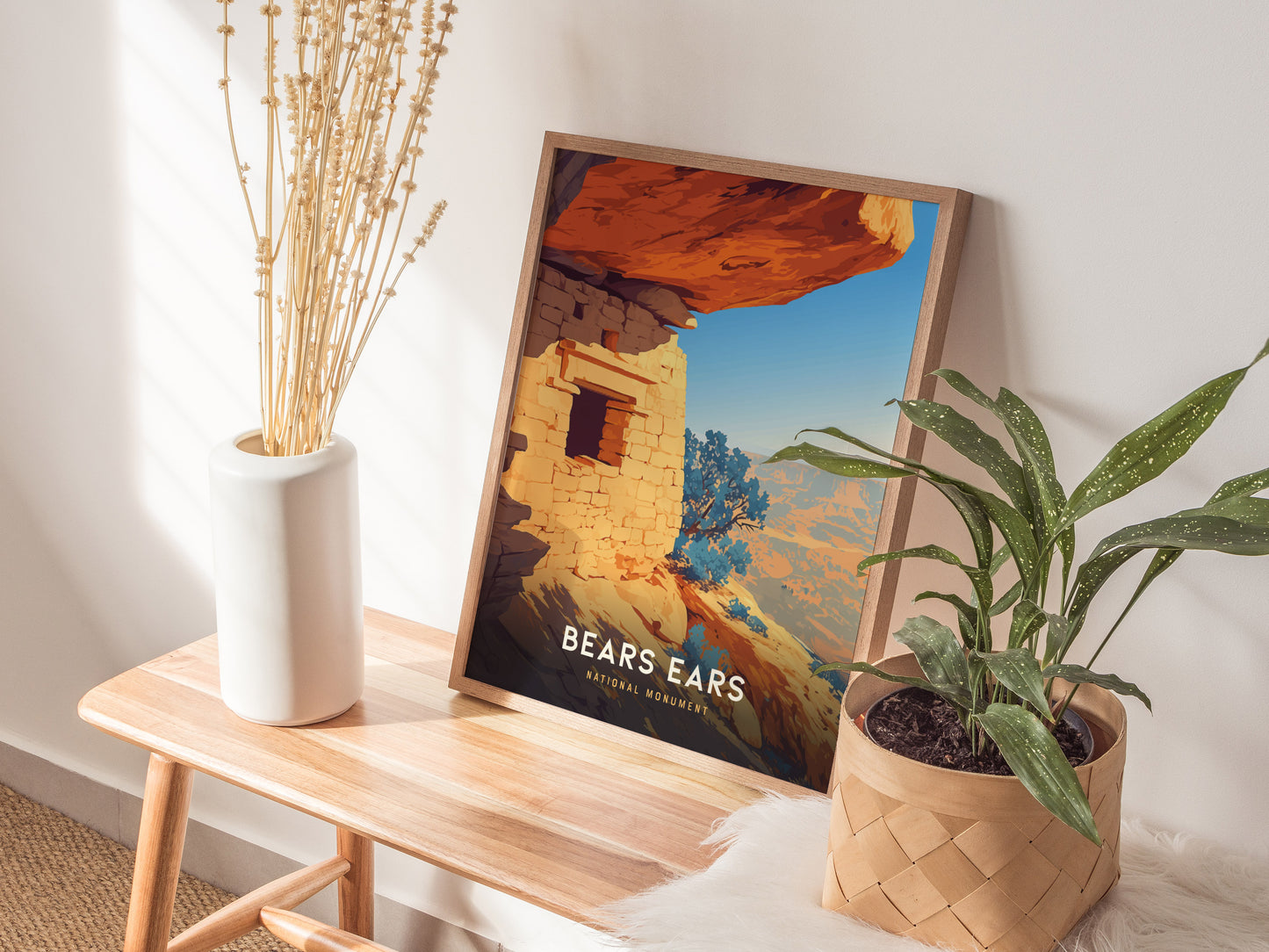 A framed Bears Ears National Monument poster leans against a wall on a wooden bench, accompanied by a white vase with dried flowers and a potted green plant.