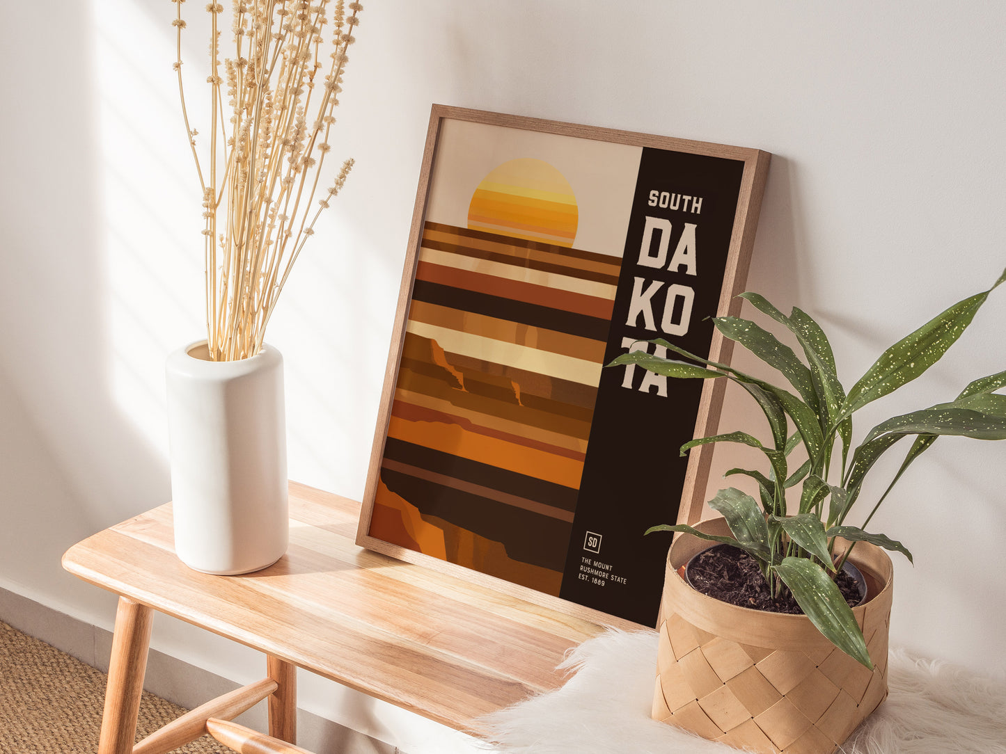 A framed South Dakota poster with geometric shapes and earthy tones is displayed on a wooden bench, flanked by a white vase with dried stems and a green potted plant.