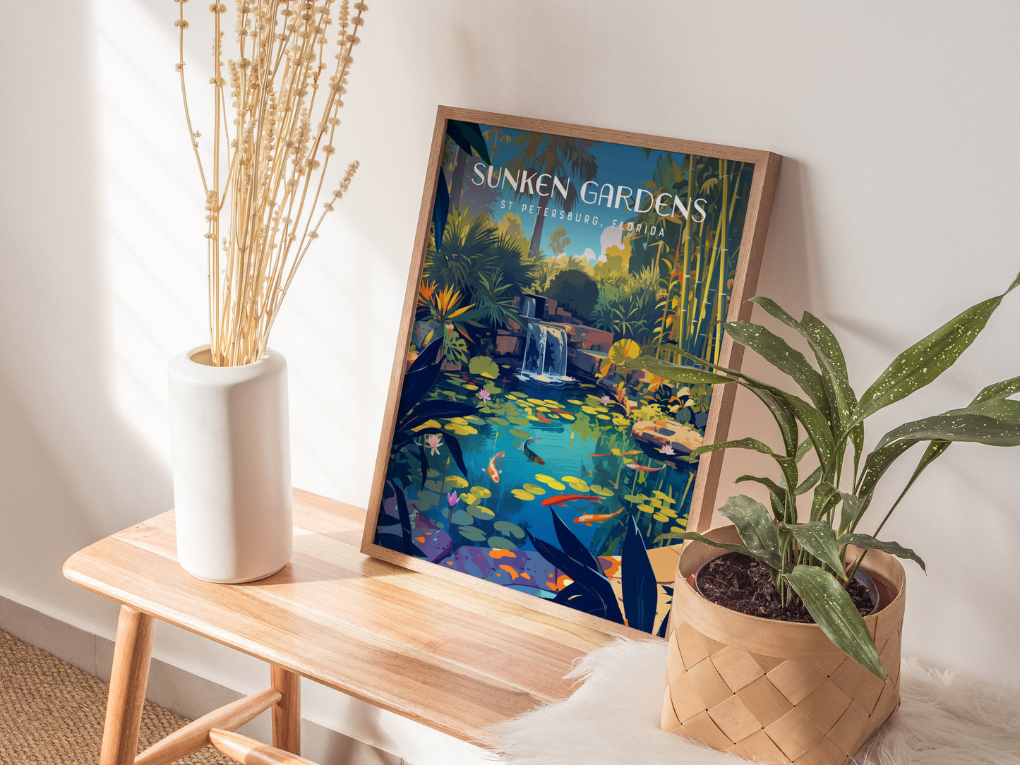 A framed Sunken Gardens St Petersburg poster depicting a serene koi pond with lush foliage and waterfalls is displayed on a wooden bench beside a vase and potted plant.