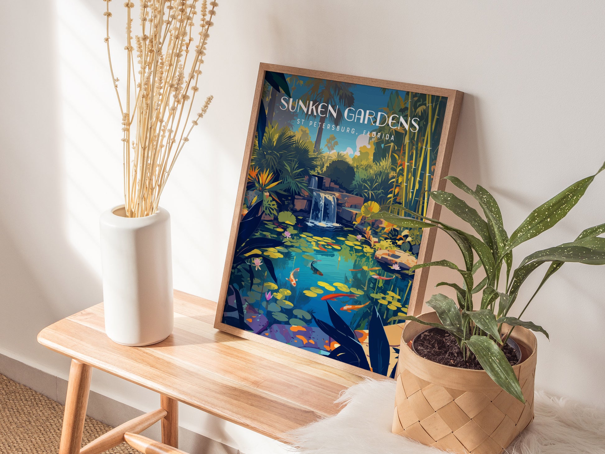 A framed Sunken Gardens St Petersburg poster depicting a serene koi pond with lush foliage and waterfalls is displayed on a wooden bench beside a vase and potted plant.