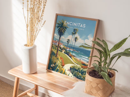A framed Encinitas, California poster depicting palm trees and a coastal view leans on a wooden bench. Nearby are a white vase with dried flowers and a potted plant.