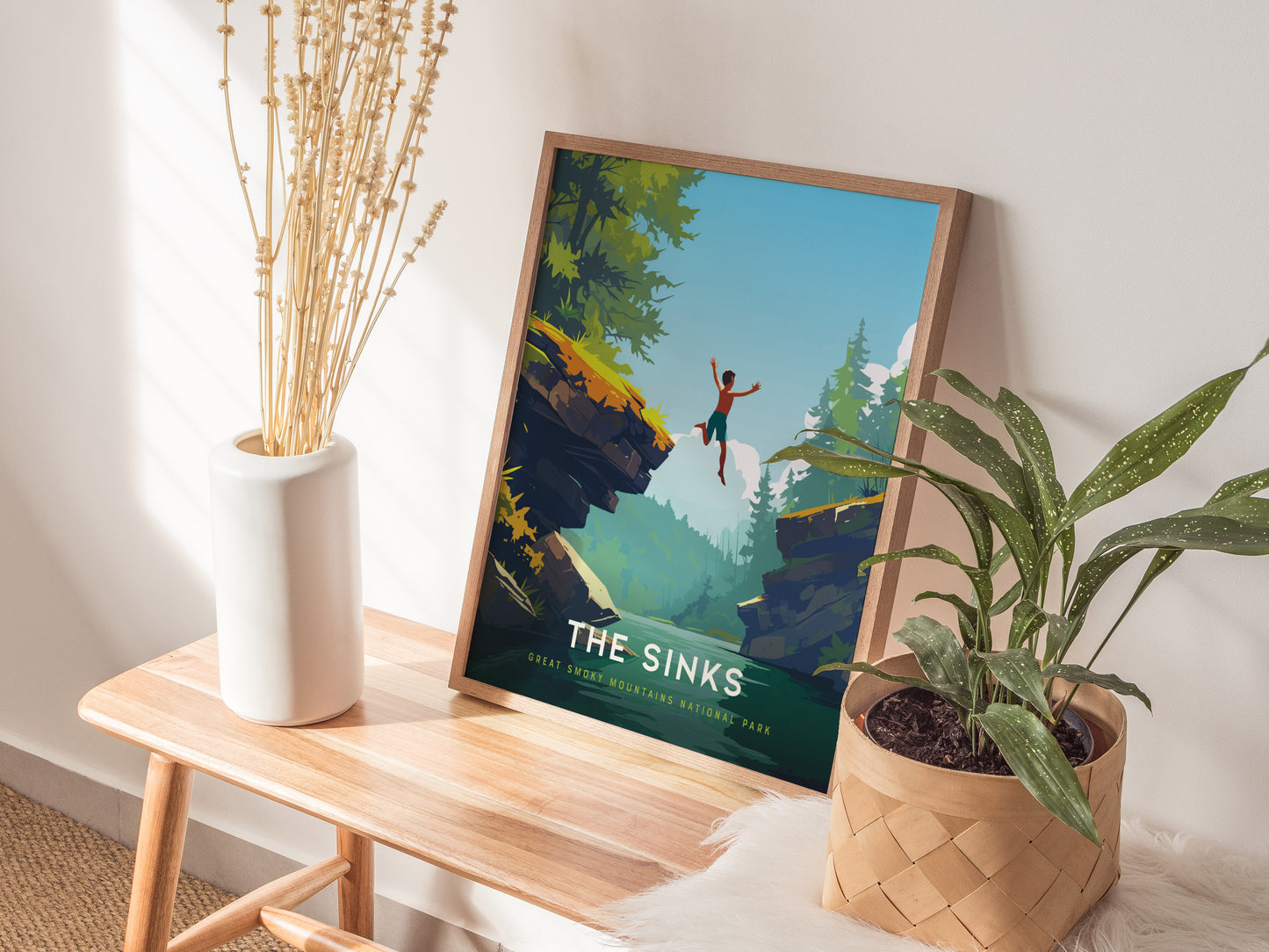 A framed poster titled The Sinks is displayed on a wooden bench, featuring an illustration of a person jumping off a cliff into a lush forest scene. A white vase with dried flowers and a potted plant are placed nearby.